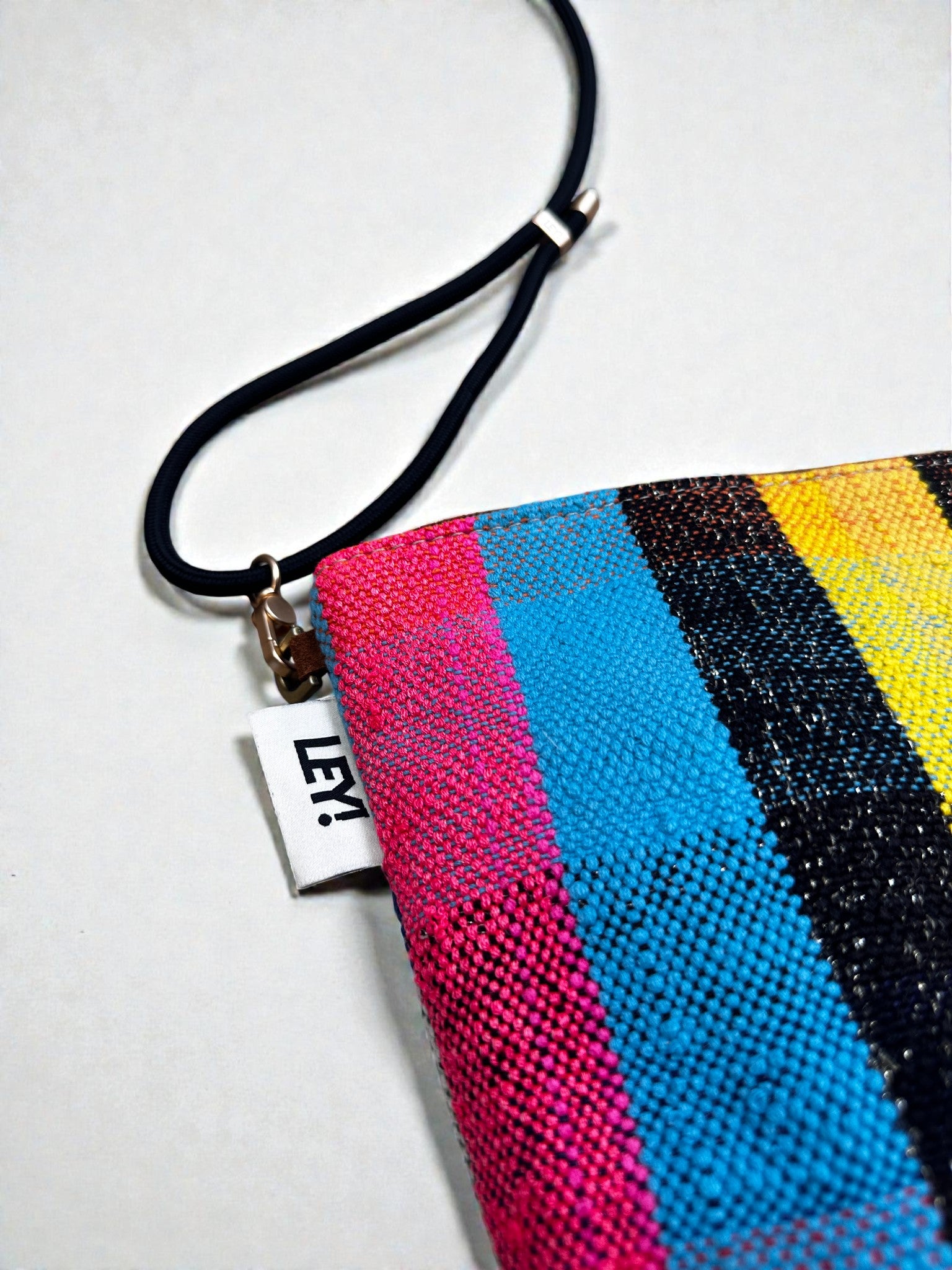 Eine Nahaufnahme der LEY! Tasche - OCULIA CLUTCH, einer farbenfrohen, handgefertigten Umhängetasche mit vertikalen Streifen in Blau, Rosa, Gelb und Schwarz, einem weißen LEY!-Schild mit schwarzem Aufdruck und einem dünnen schwarzen Riemen mit Metallverschluss.