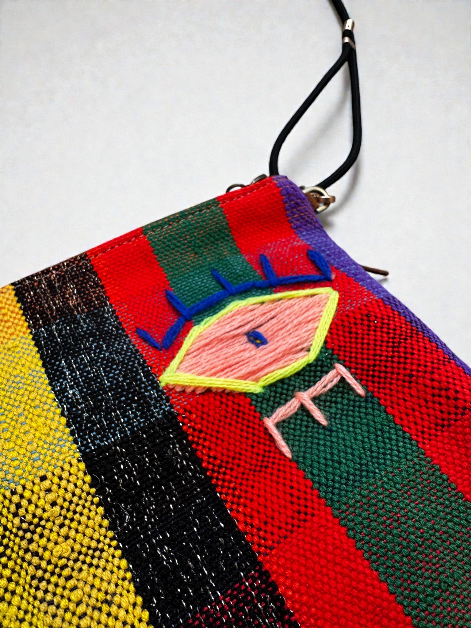 Die LEY! Tasche - OCULIA CLUTCH von LEY! ist eine bunte, gewebte Clutch mit vertikalen Streifen in Rot, Grün, Gelb und Schwarz. In der Mitte ist ein abstraktes Auge aufgestickt und an der oberen Ecke ist ein schwarzer Riemen befestigt.