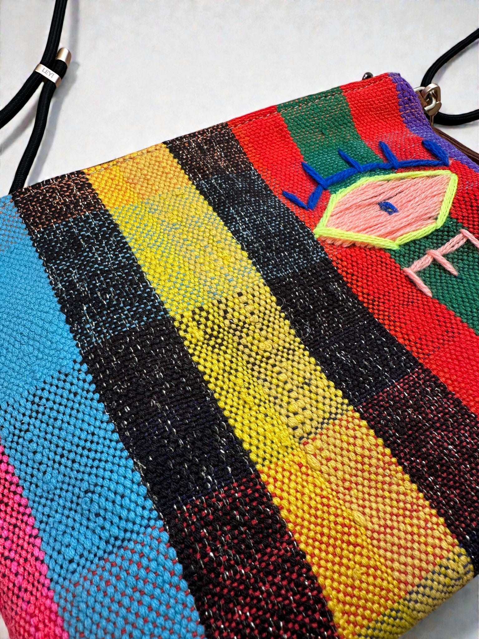 Die LEY! Tasche - OCULIA CLUTCH von LEY! ist eine handgefertigte Umhängetasche aus Vintage-Stoff mit vertikalen Streifen in Blau, Schwarz, Gelb und Rot sowie abstraktem Augenmotiv in Pink, Blau und Grün am oberen rechten Rand.