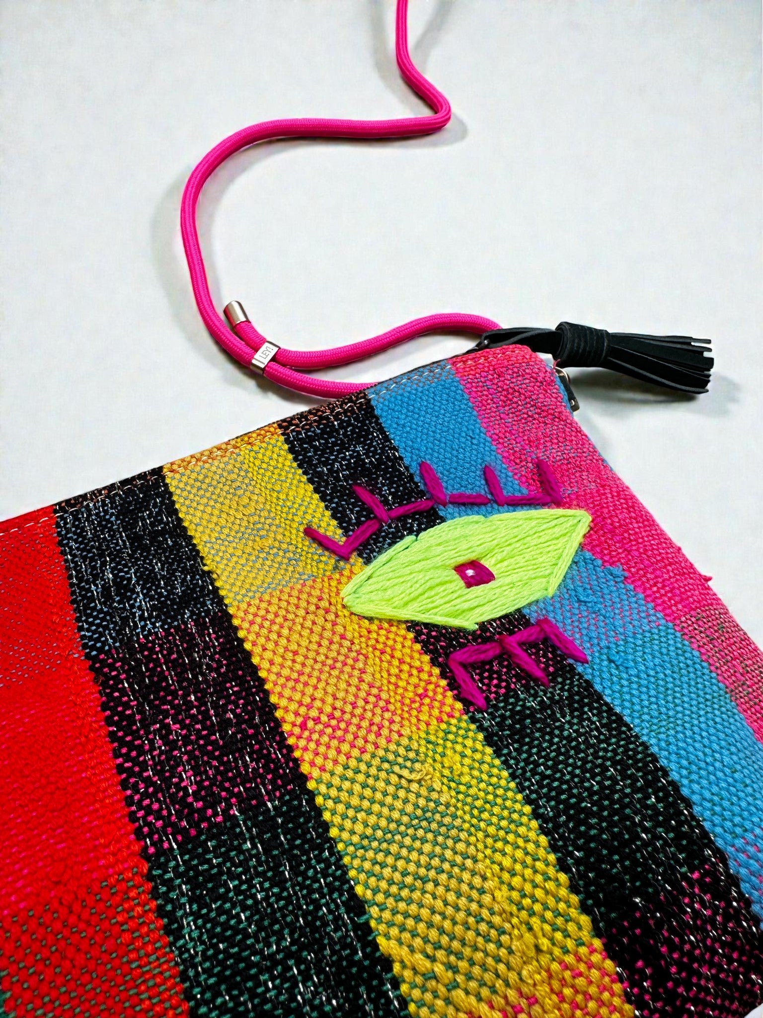 Die LEY! Tasche - OCULIA CLUTCH von LEY! ist eine farbenfrohe, gewebte Tasche mit roten, gelben, blauen und schwarzen Streifen, einem gestickten Auge auf Vintage-Stoff, sowie einer pinken Reißverschluss-Kordel und einer schwarzen Quaste.