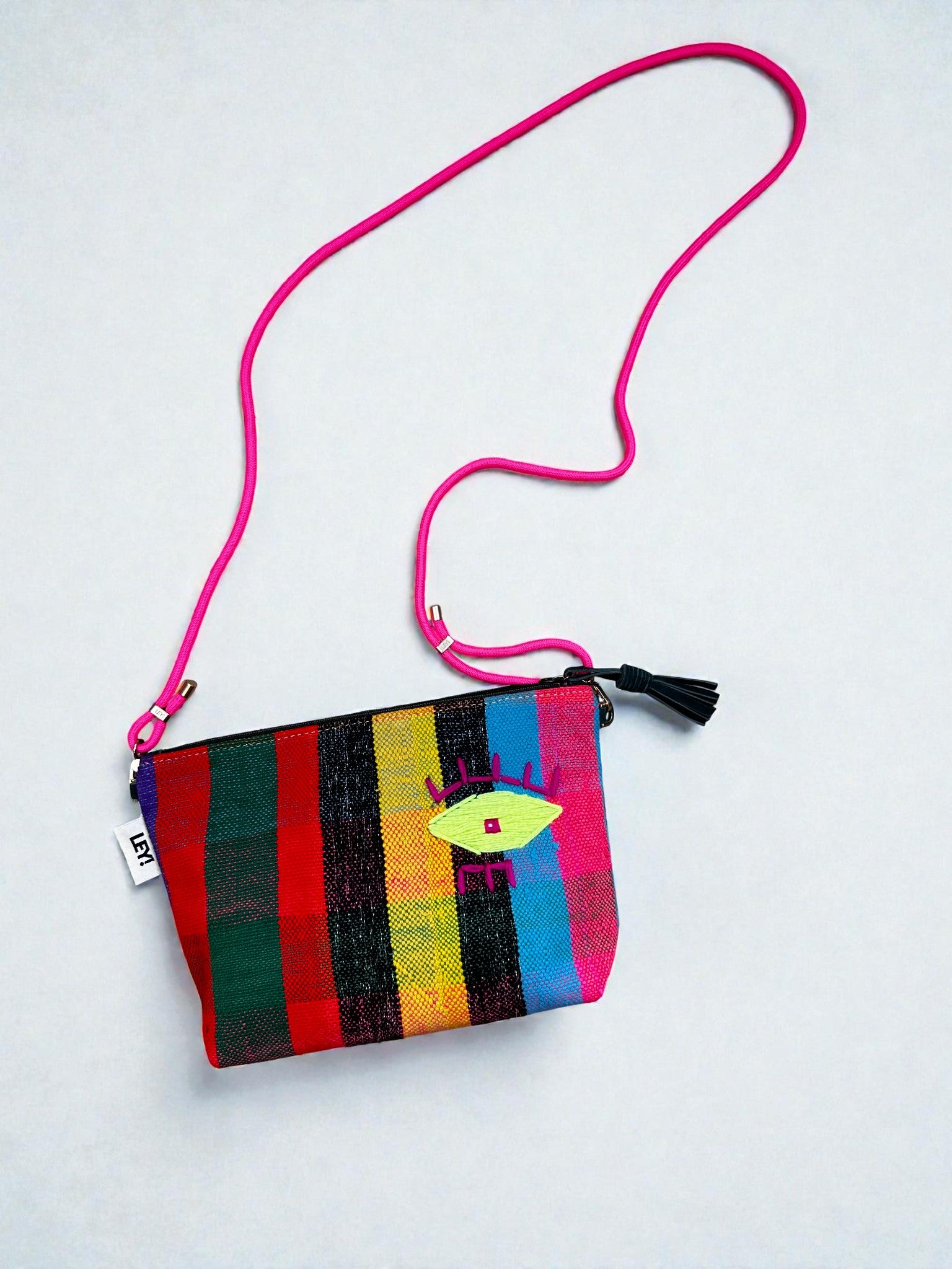 Die LEY! Tasche - OCULIA CLUTCH ist eine kleine, mehrfarbig gestreifte Clutch mit rosafarbenem Riemen, schwarzer Quaste, leuchtend gelbem Augenmotiv mit rosafarbenen und violetten Akzenten und einem seitlichen Anhänger mit der Aufschrift "LEY!".