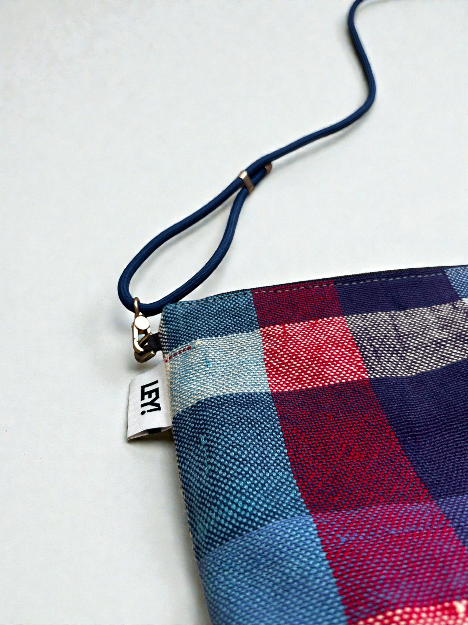 Eine Nahaufnahme der bunten, karierten LEY! Tasche - OCULIA CLUTCH von LEY!, handgefertigt aus Vintage-Stoff mit einem blauen Riemen und einem kleinen weißen "LEM"-Label auf hellem Grund.