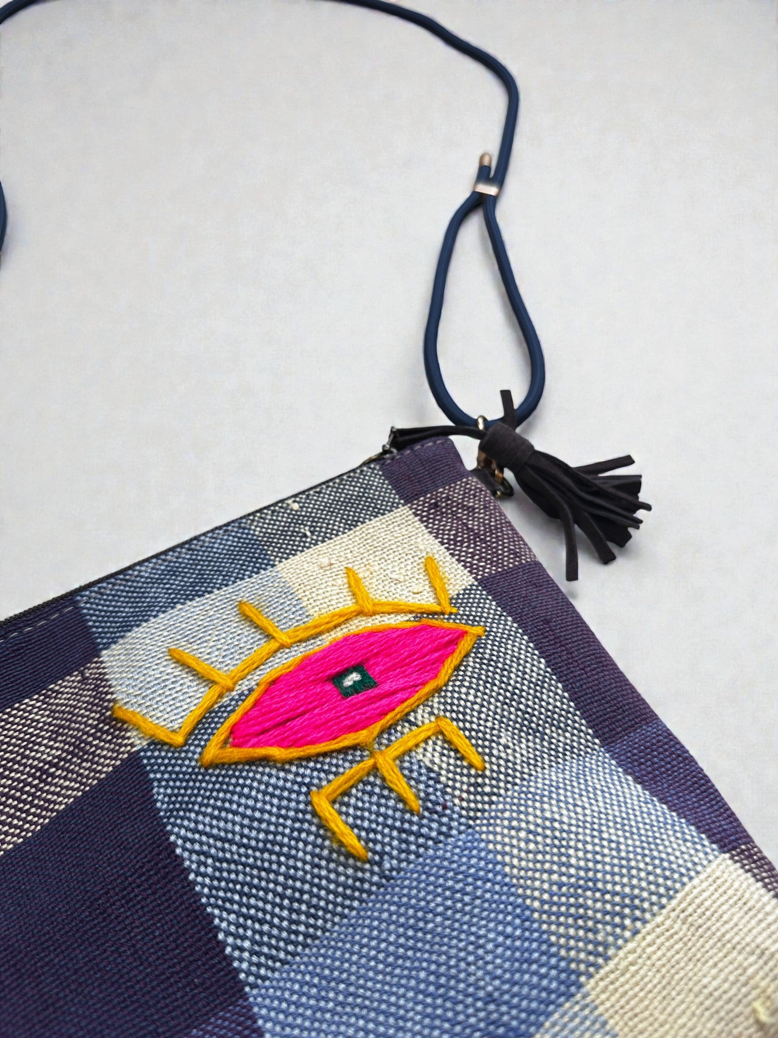 Die LEY! Tasche - OCULIA CLUTCH von LEY! ist eine handgefertigte Umhängetasche aus Vintage-Stoff mit schwarzem Riemen, Quaste und gelb, pink und blau gesticktem Augenmotiv auf der Vorderseite.