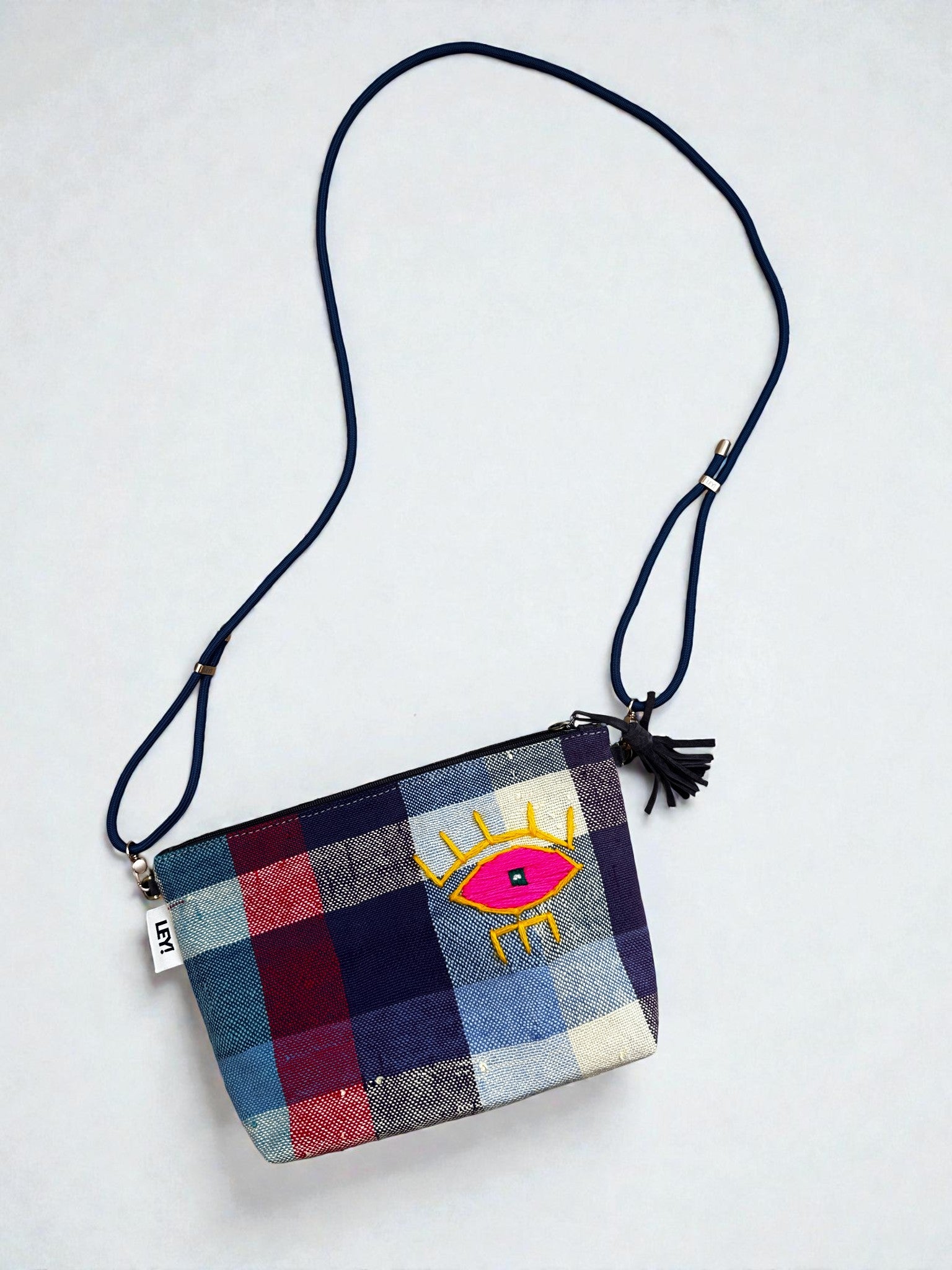Die LEY! Tasche - OCULIA CLUTCH von LEY! ist eine handgefertigte Umhängetasche aus Vintage-Stoff mit blau-rotem Karomuster, gelb-pinker Oculia-Augenstickerei, blauem Riemen und schwarzem Quastenanhänger.