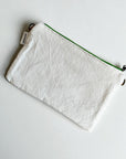Eine rechteckige weiße Stofftasche mit einem neongrünen Reißverschluss und einem kleinen weißen Anhänger - die LEY! Tasche - Mona neongrün - 20x30cm ruht elegant auf einer schlichten weißen Oberfläche.