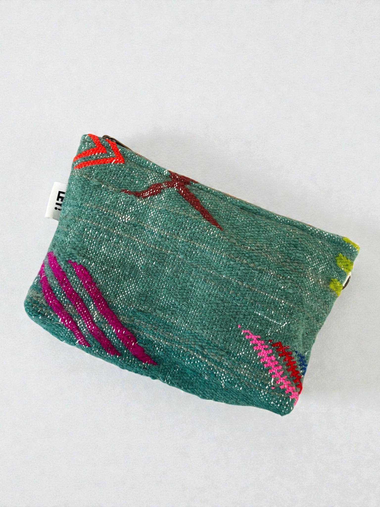 Die LEY! Tasche - Colorful Line (20 x 30 cm) ist eine kleine tealfarbene Tasche mit roten, rosa und gelben Stickmustern, handgefertigt in Marrakesch Handarbeit. Sie hat ein weißes Seitenschild und wird auf einer hellen Fläche ausgestellt.