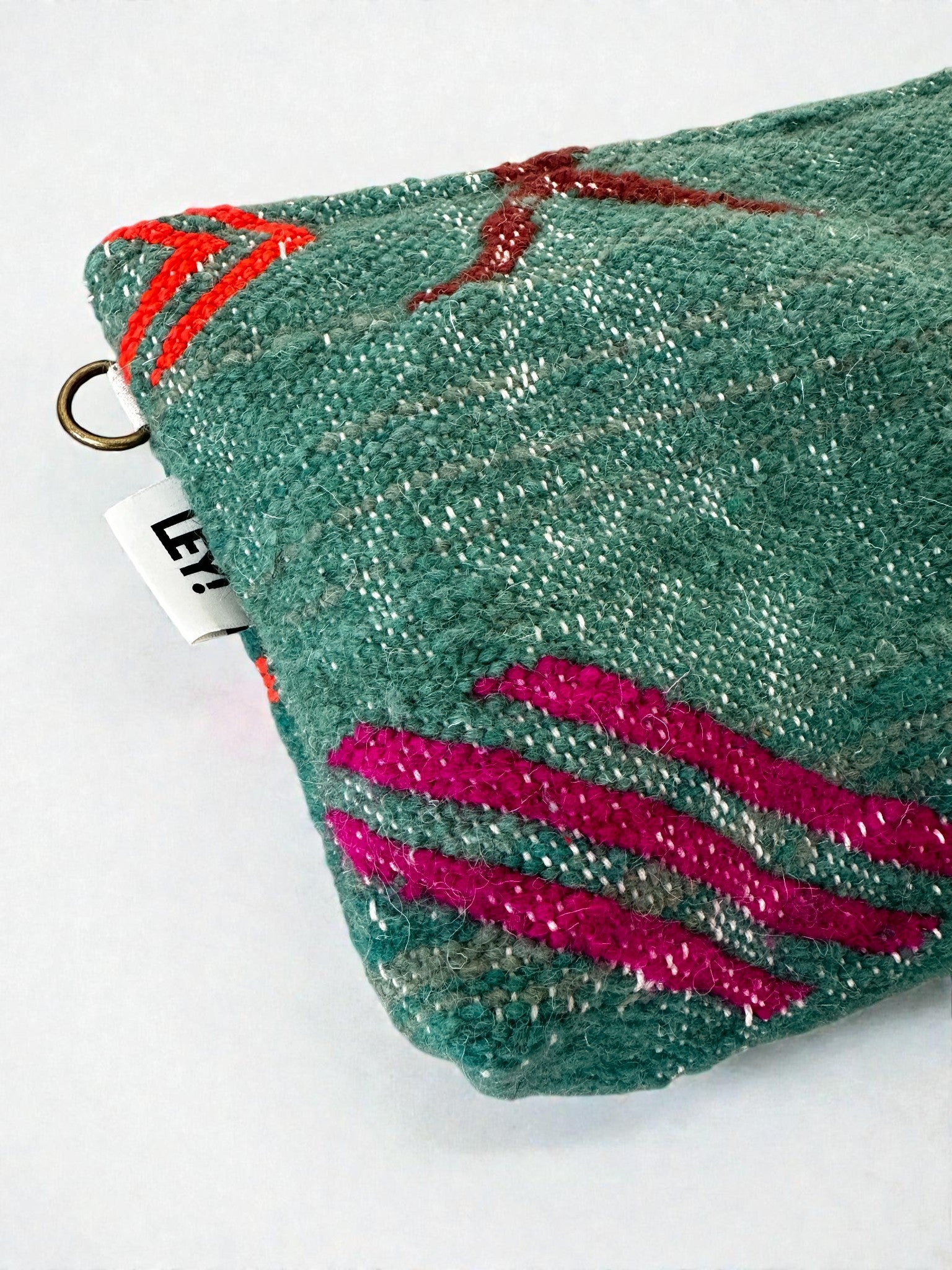 Eine Nahaufnahme der LEY! Tasche - Colorful Line - 20 x 30 cm aus grüner Schurwolle mit rosa und roter Stickerei, einem weißen Anhänger mit schwarzem Text und einem Messingring in der Ecke. Handgefertigt in Marrakesch, vor weißem Hintergrund.