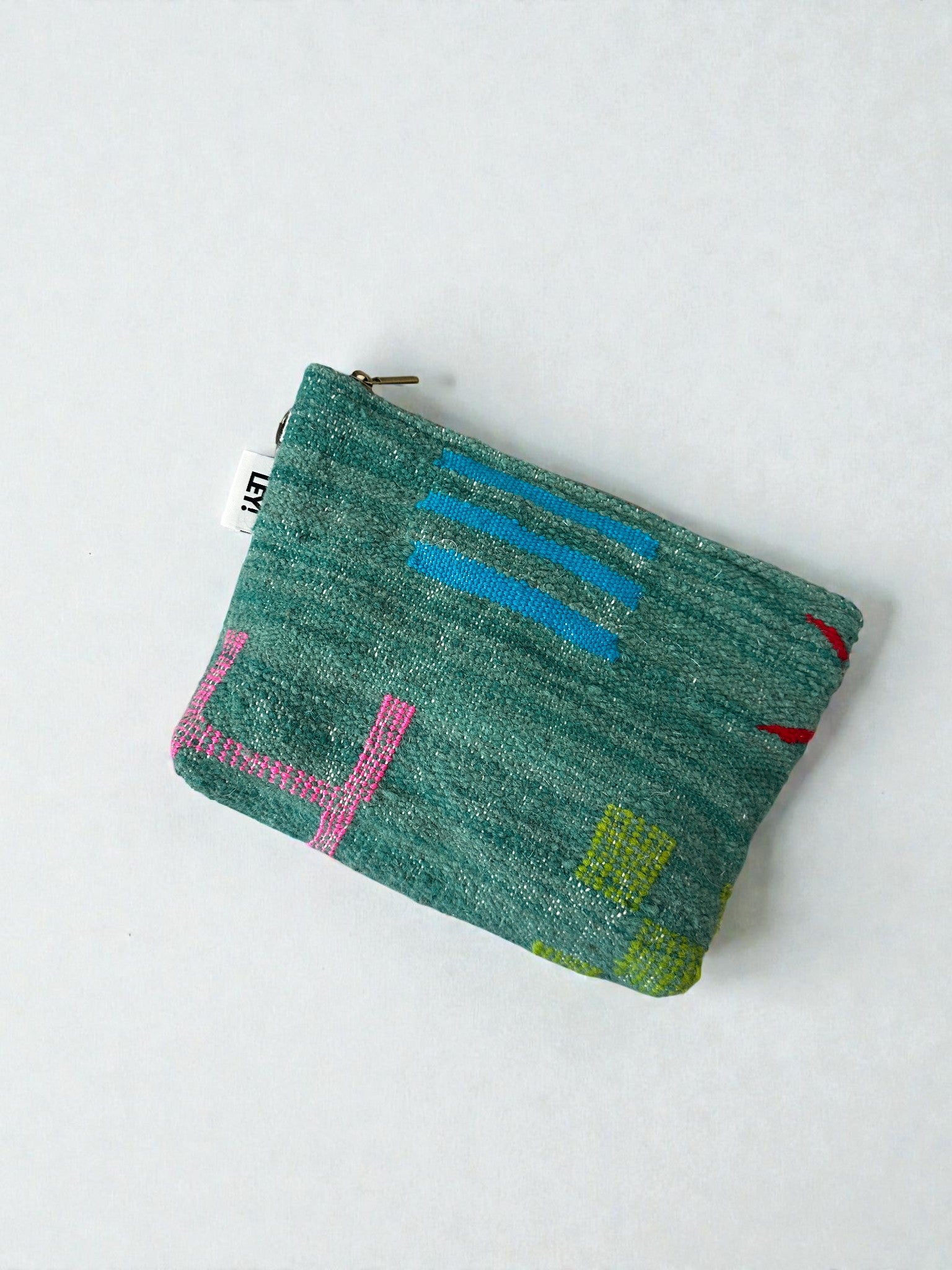 Die Tasche - Colorful Line - 20 x 30 cm ist eine kleine, rechteckige, grüne Stofftasche von LEY! mit abstrakten, gestickten blauen, rosa und gelben Mustern auf hellem Grund und einem weißen Etikett mit schwarzer Schrift in der Nähe des Reißverschlusses.