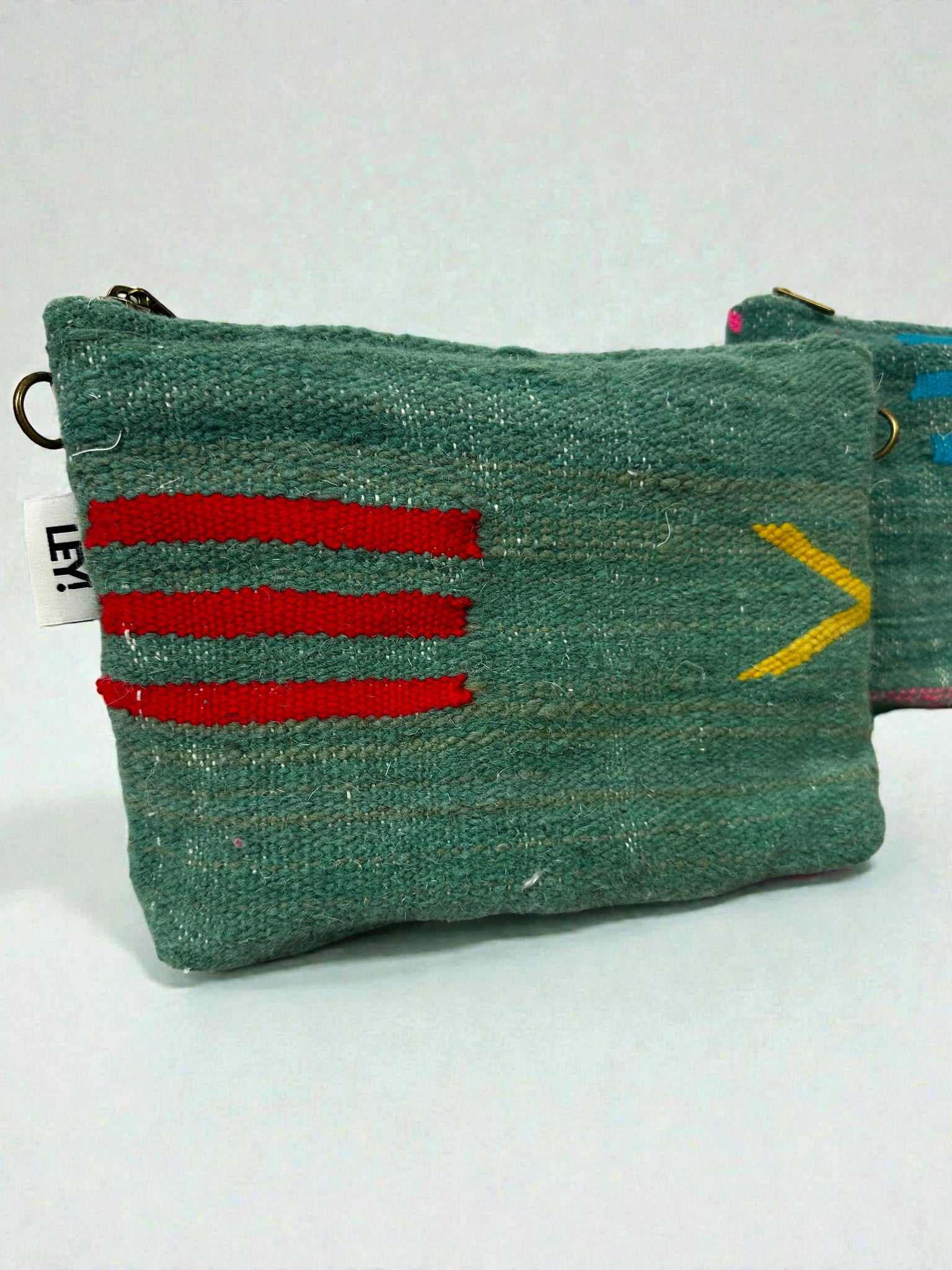Eine kleine grüne gewebte LEY! Tasche - Colorful Line (20 x 30 cm) mit einem Reißverschluss, drei roten Akzenten und einem gelben Chevron. Auf der linken Seite befindet sich ein weißes Etikett mit schwarzer Schrift; eine weitere ähnliche Tasche ist dahinter teilweise sichtbar.
