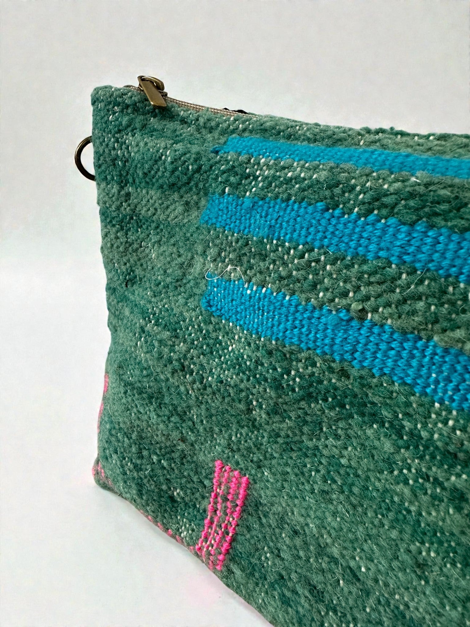Eine Nahaufnahme der LEY! Tasche - Colorful Line (20 x 30 cm) zeigt strukturierte grüne Schurwolle mit blauen und rosafarbenen Flechtmustern, einen Reißverschluss oben und einen Metallring. Der Hintergrund ist schlicht und hell - Handarbeit im Marrakesch-Stil.