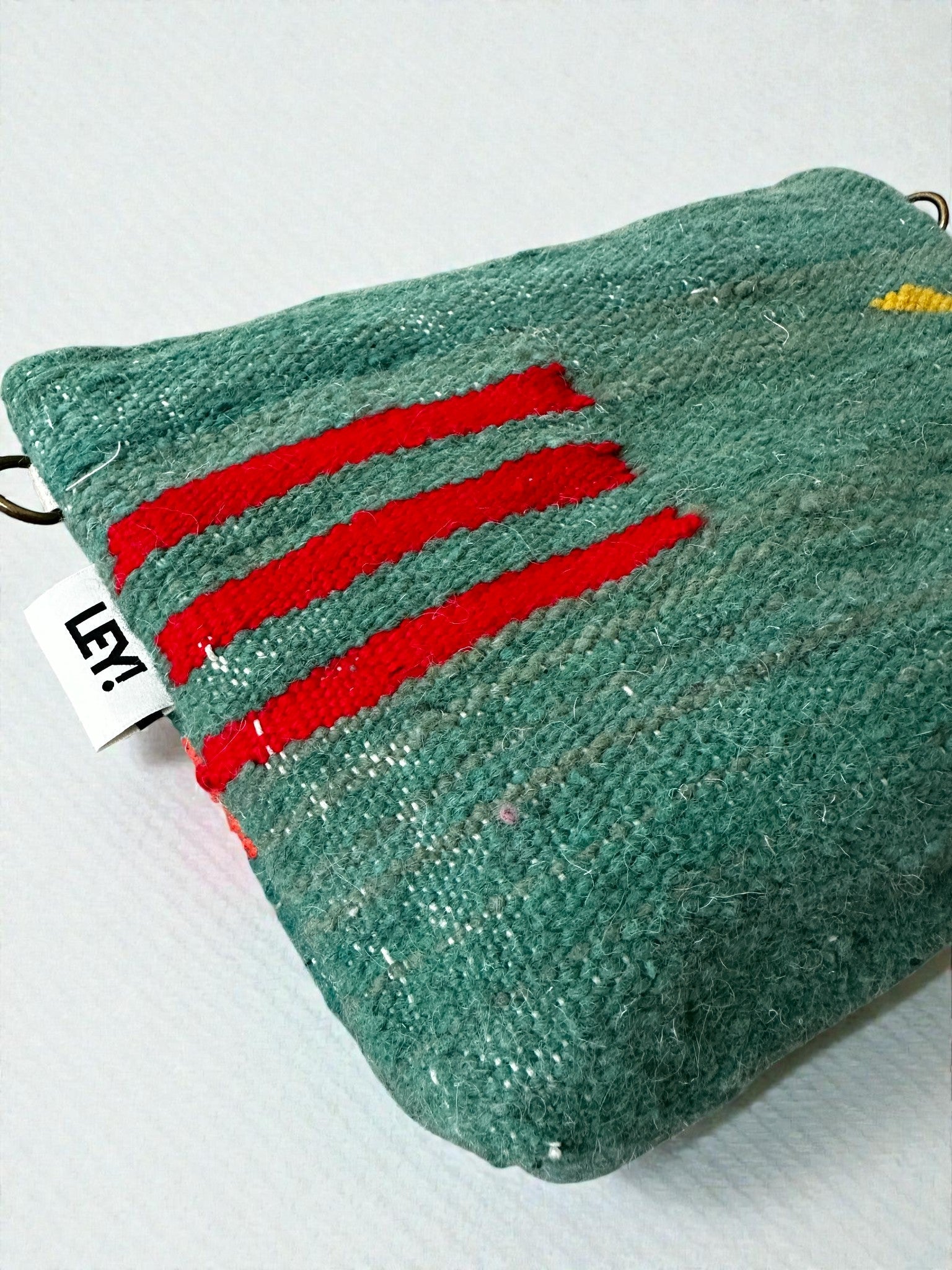 Eine strukturierte grüne Schurwolle LEY! Tasche - Colorful Line (20 x 30 cm) zeigt drei kräftige rote Linien, einen kleinen gelben Aufnäher und ein seitliches weißes Etikett mit schwarzem Text. Diese handgefertigte Tasche ruht auf einer leicht gemusterten Oberfläche.