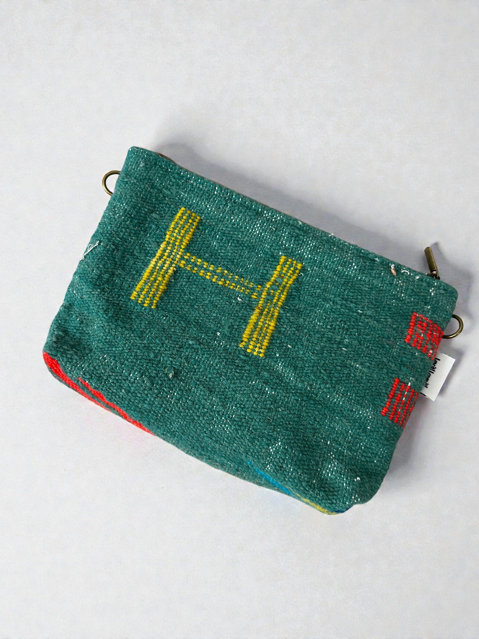 Eine kleine grüne LEY! Tasche - Colorful Line - 20 x 30 cm große Reißverschlusstasche mit einem gelben "H" auf der Vorderseite, roten Seitennähten und einem Metallring an einer Ecke, auf hellem Hintergrund.