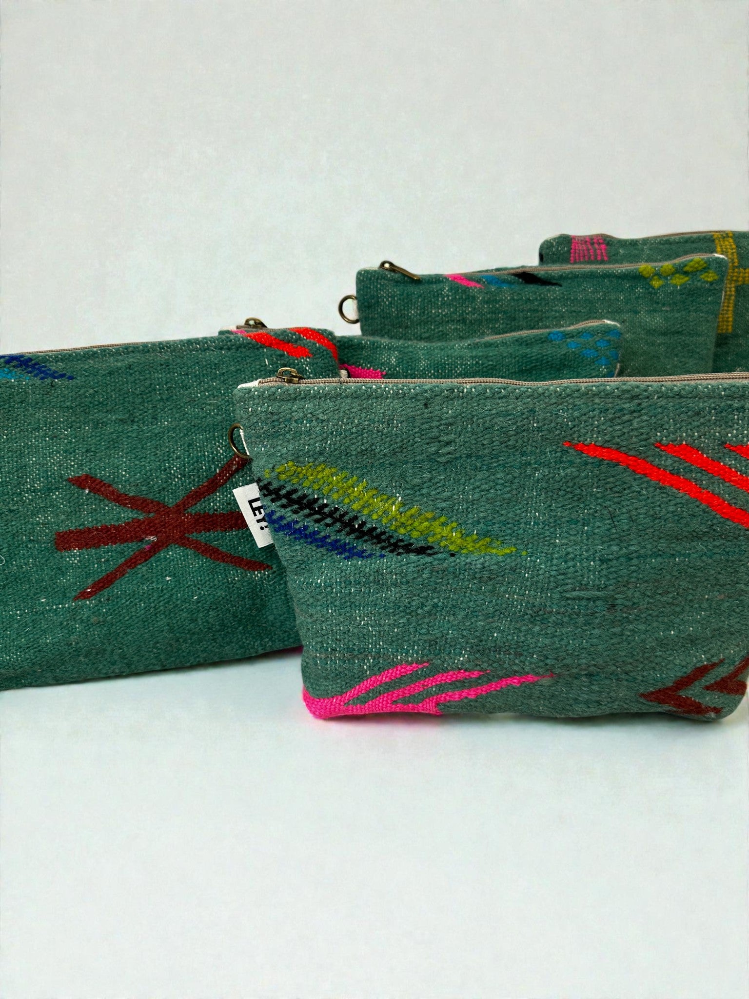 Vier grüne LEY! Tasche - Colorful Line (20 x 30 cm) mit roter, blauer, pinker und gelber Stickerei werden vor einem einfarbigen Hintergrund gezeigt. Jede hat einen Reißverschluss und zeigt exquisite Handarbeit aus Marrakesch.