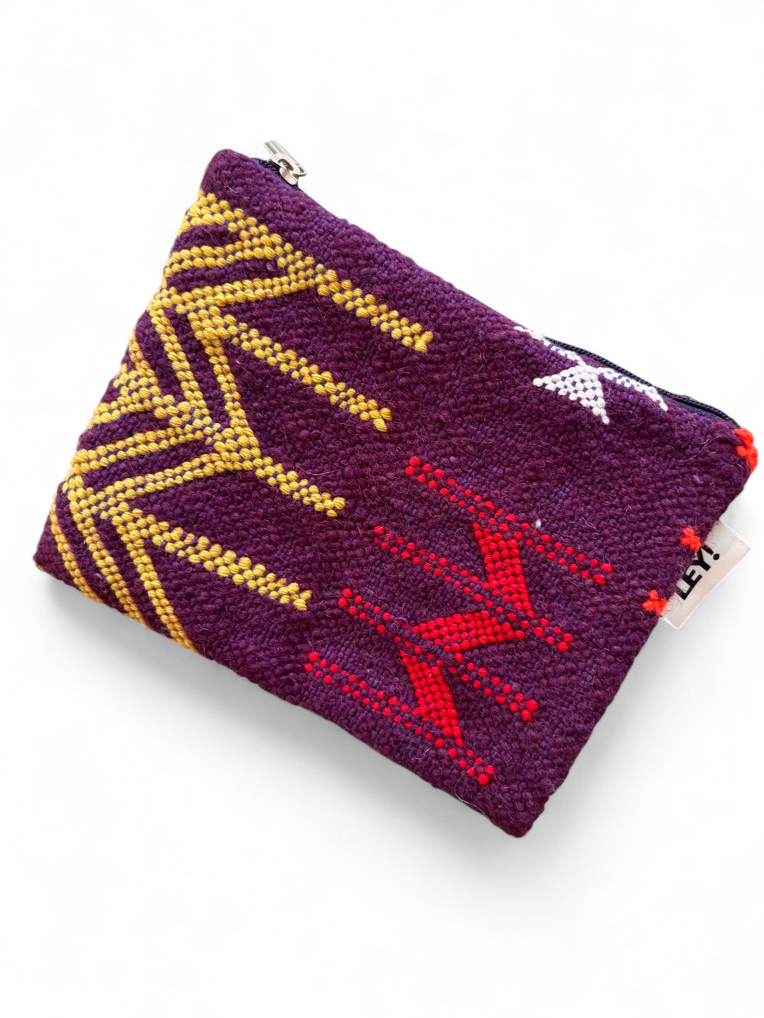 Die LEY! Tasche - Colorful Line (15x20cm) ist aus Schurwolle in Lila mit gelb-roten geometrischen Mustern, weißem Stern-Design, Reißverschluss oben und kleinem weißen Tag mit schwarzer Aufschrift. Handgefertigt in Marrakesch.