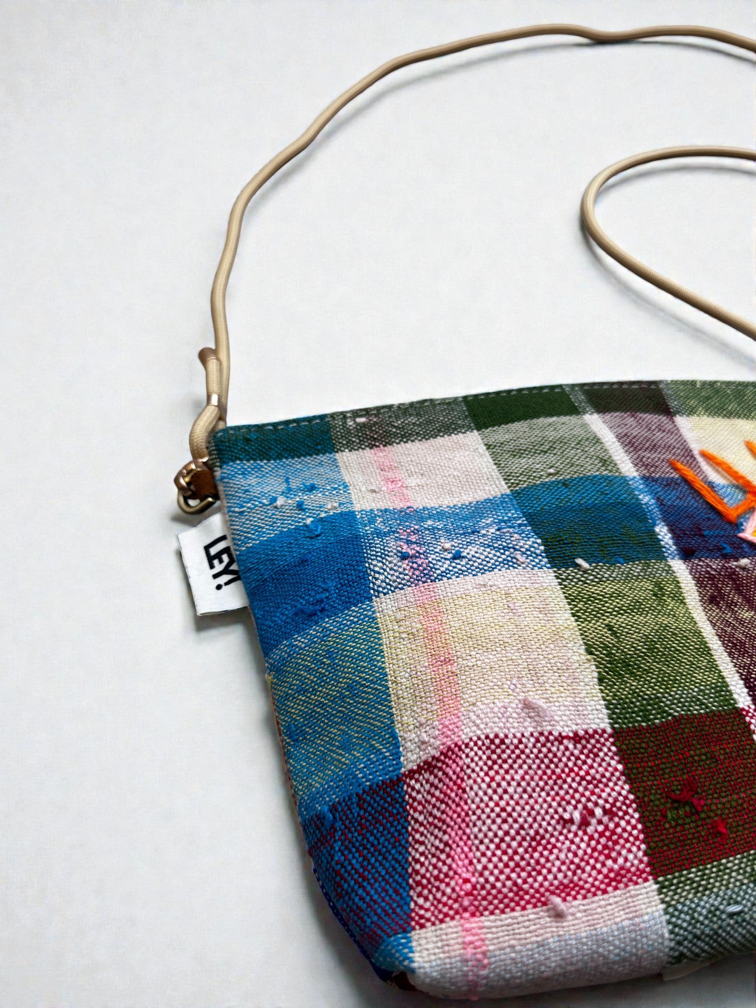 Eine Nahaufnahme der LEY! Tasche - OCULIA CLUTCH von LEY!, einer bunt karierten Tasche mit beigem Riemen, die auf einer weißen Fläche abgebildet ist. Ein weißes Seitenetikett mit schwarzem Text ist sichtbar.
