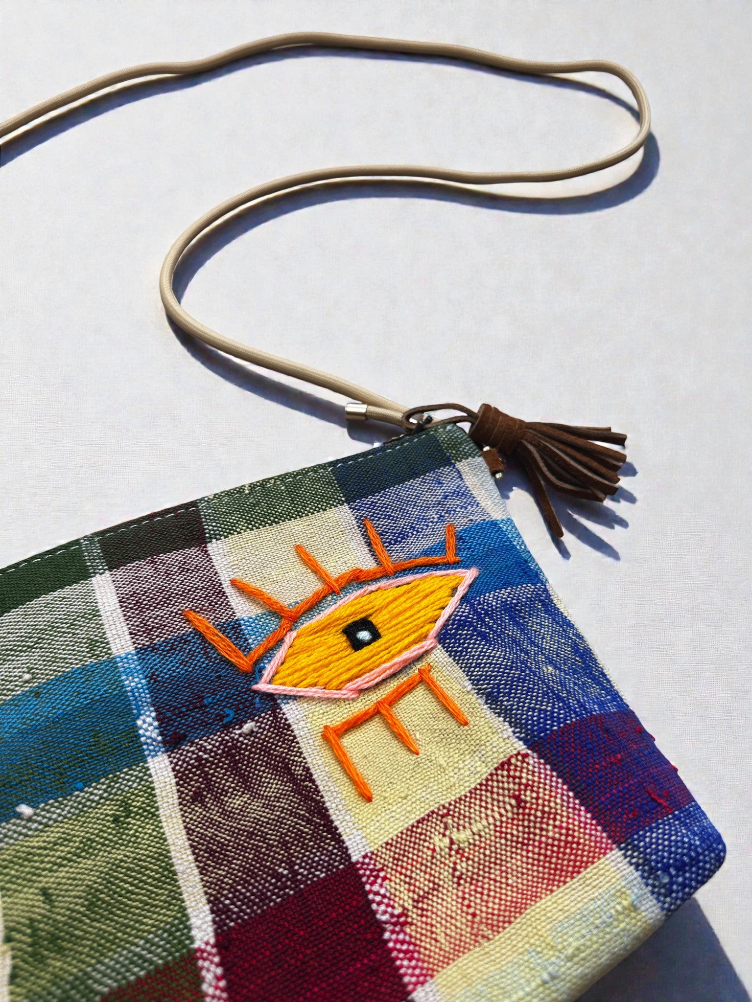 Die LEY! Tasche - OCULIA CLUTCH von LEY! hat ein buntes Karomuster, einen braunen Riemen, eine braune Quaste und ein gesticktes gelbes Auge mit orangefarbenen Strahlen auf der Vorderseite, das stilvoll auf einer hellen Oberfläche ruht.