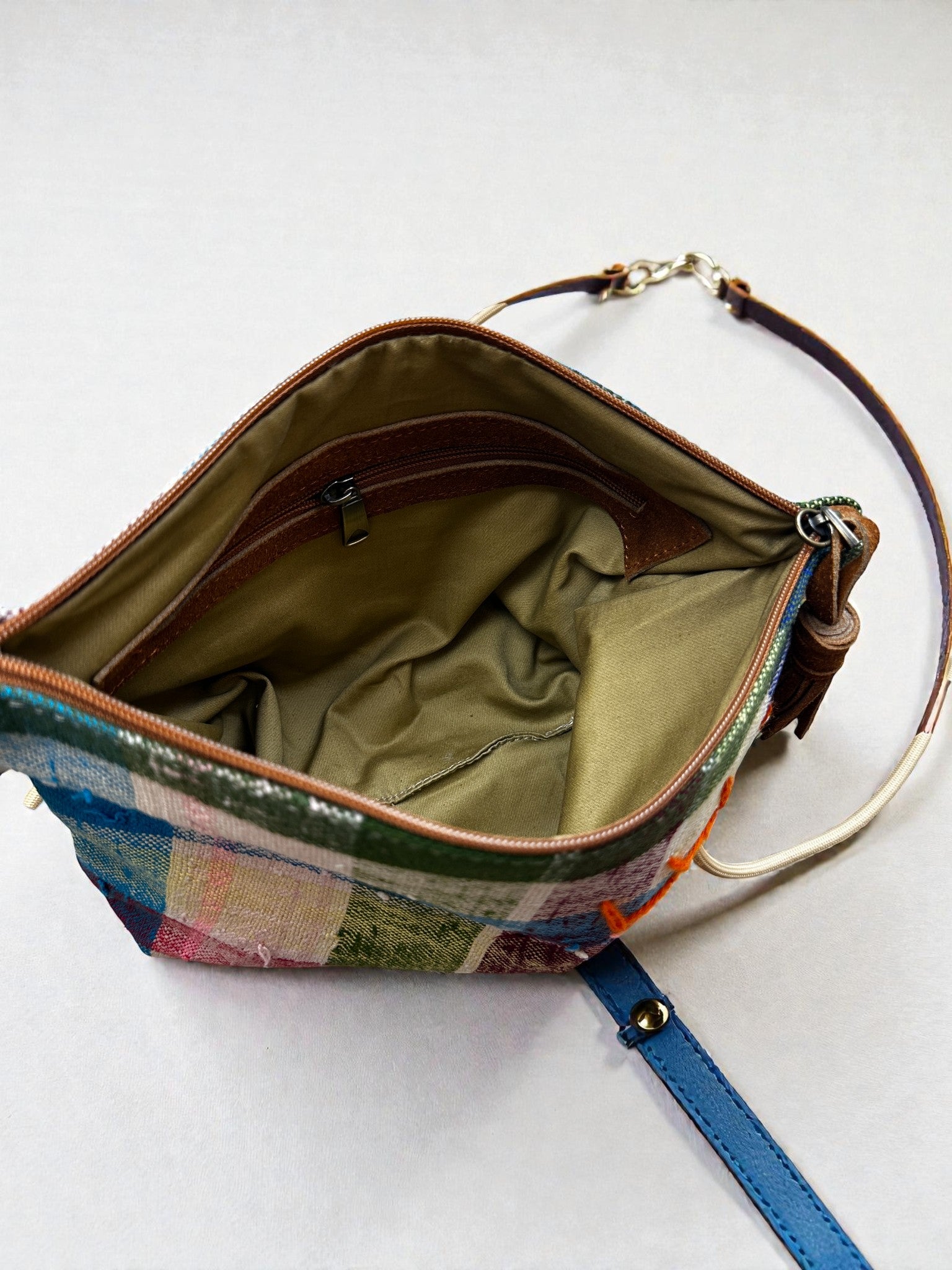 Eine Draufsicht auf die LEY! Tasche - OCULIA CLUTCH von LEY! zeigt eine handgefertigte Umhängetasche mit braunem Reißverschluss, beigefarbenem Stofffutter, Innentasche und teils sichtbarem abnehmbarem blau-braunen Träger.