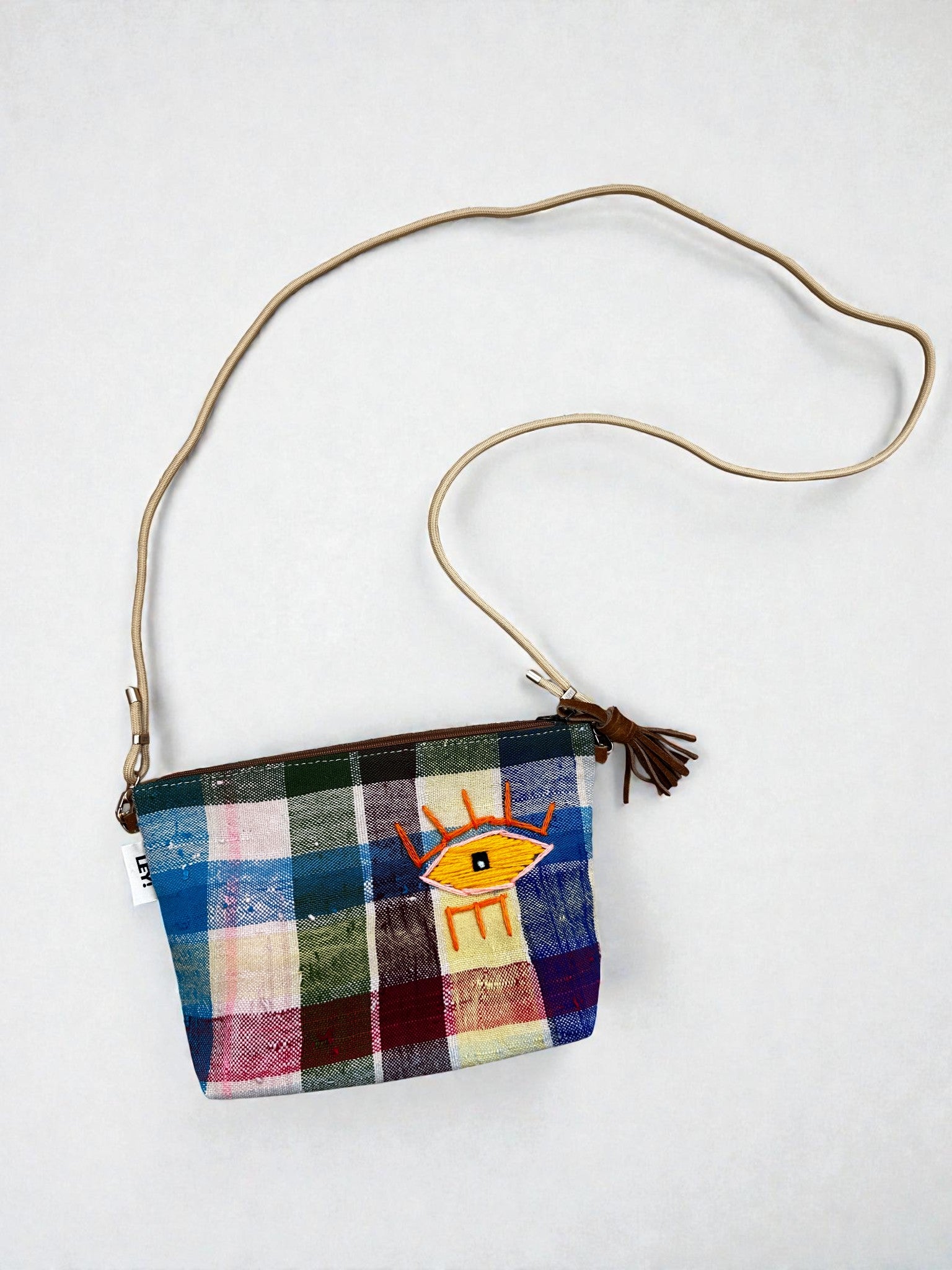 Die LEY! Tasche - OCULIA CLUTCH von LEY! ist eine handgefertigte Umhängetasche aus buntem Vintage-Stoff mit tanfarbenem Riemen, gesticktem gelben Auge vorne, orangefarbenen Wimpern, Reißverschluss und braunem Quasten-Detail.
