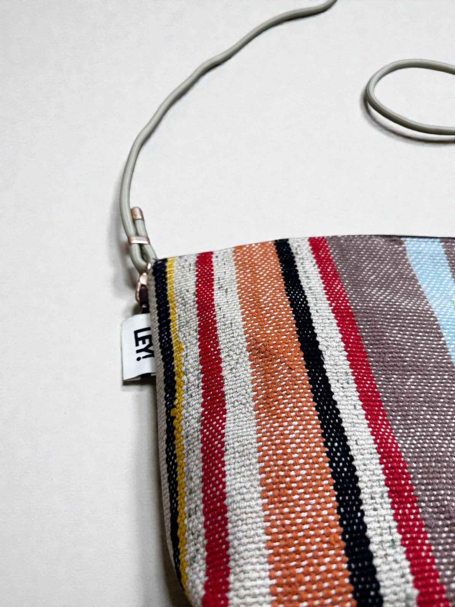 Eine Nahaufnahme der LEY! Tasche - OCULIA CLUTCH von LEY!, einer handgefertigten Clutch mit leuchtend roten, orangen, gelben, schwarzen, braunen und blauen Längsstreifen, einem schmalen Riemen und einem kleinen weißen "LE17"-Anhänger.