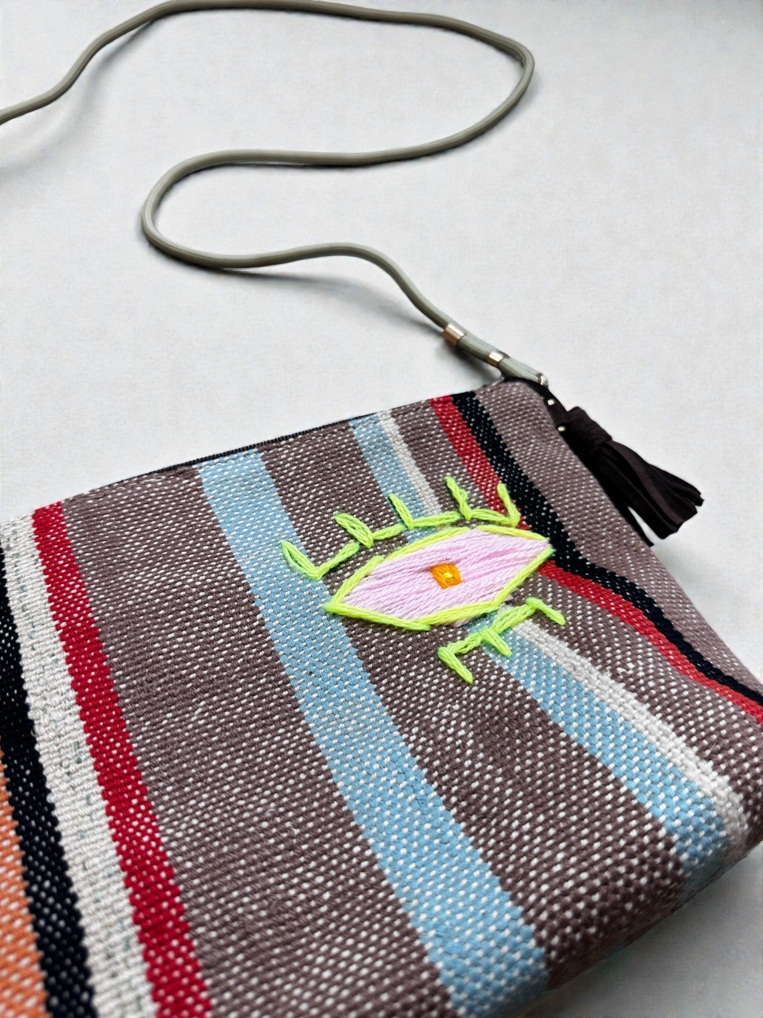 Die LEY! Tasche - OCULIA CLUTCH von LEY! ist eine kleine gewebte Clutch mit roten, blauen, braunen und grauen Streifen, einem neongrünen gestickten Auge, einem grauen Riemen mit Quaste und Vintage-Stoff-Flair für jedes Outfit.