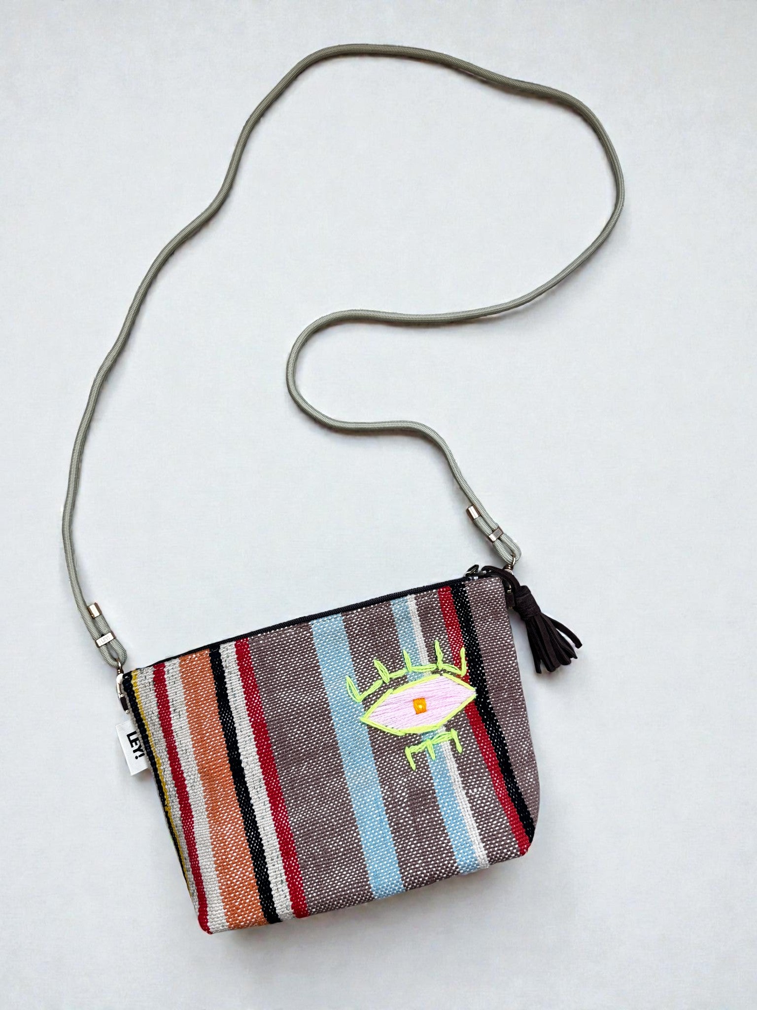 Die LEY! Tasche - OCULIA CLUTCH von LEY! ist eine kleine Crossbody-Tasche mit bunt gestreiftem Stoff, gesticktem Augenmotiv, Quasten-Reißverschluss und einem dünnen grauen Riemen.