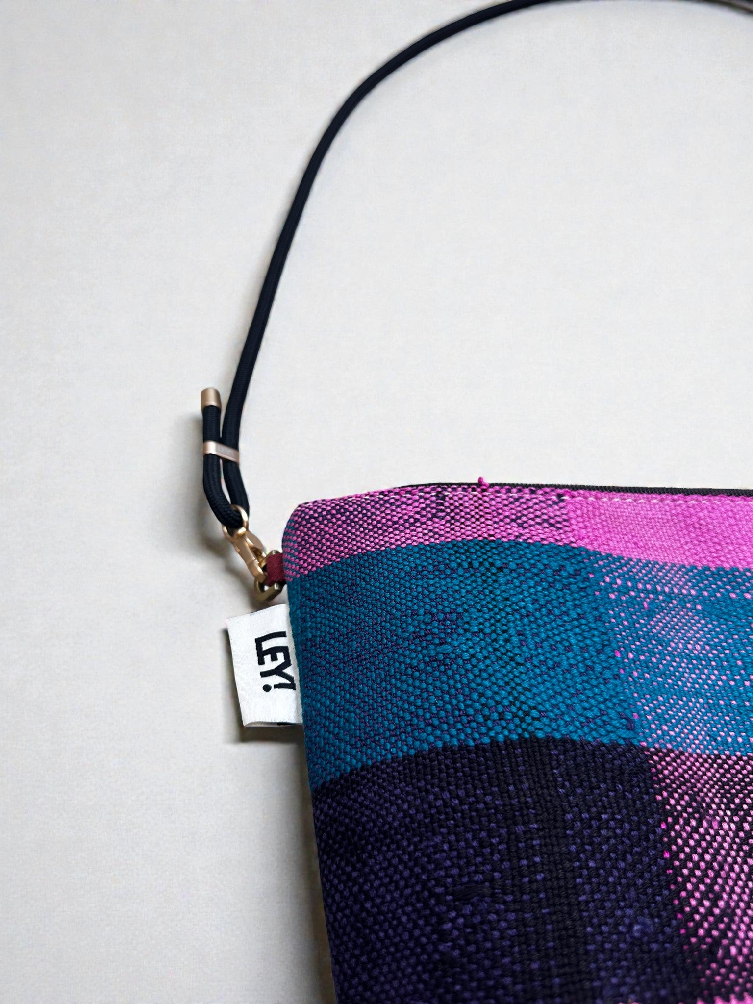 Nahaufnahme der LEY! Tasche - OCULIA CLUTCH von LEY!: Eine farbenfrohe, handgemachte Umhängetasche aus Vintage-Stoff mit schwarzem Gurt und "LEY!"-Tag in Pink-, Blau- und Lilatönen vor hellem Hintergrund.