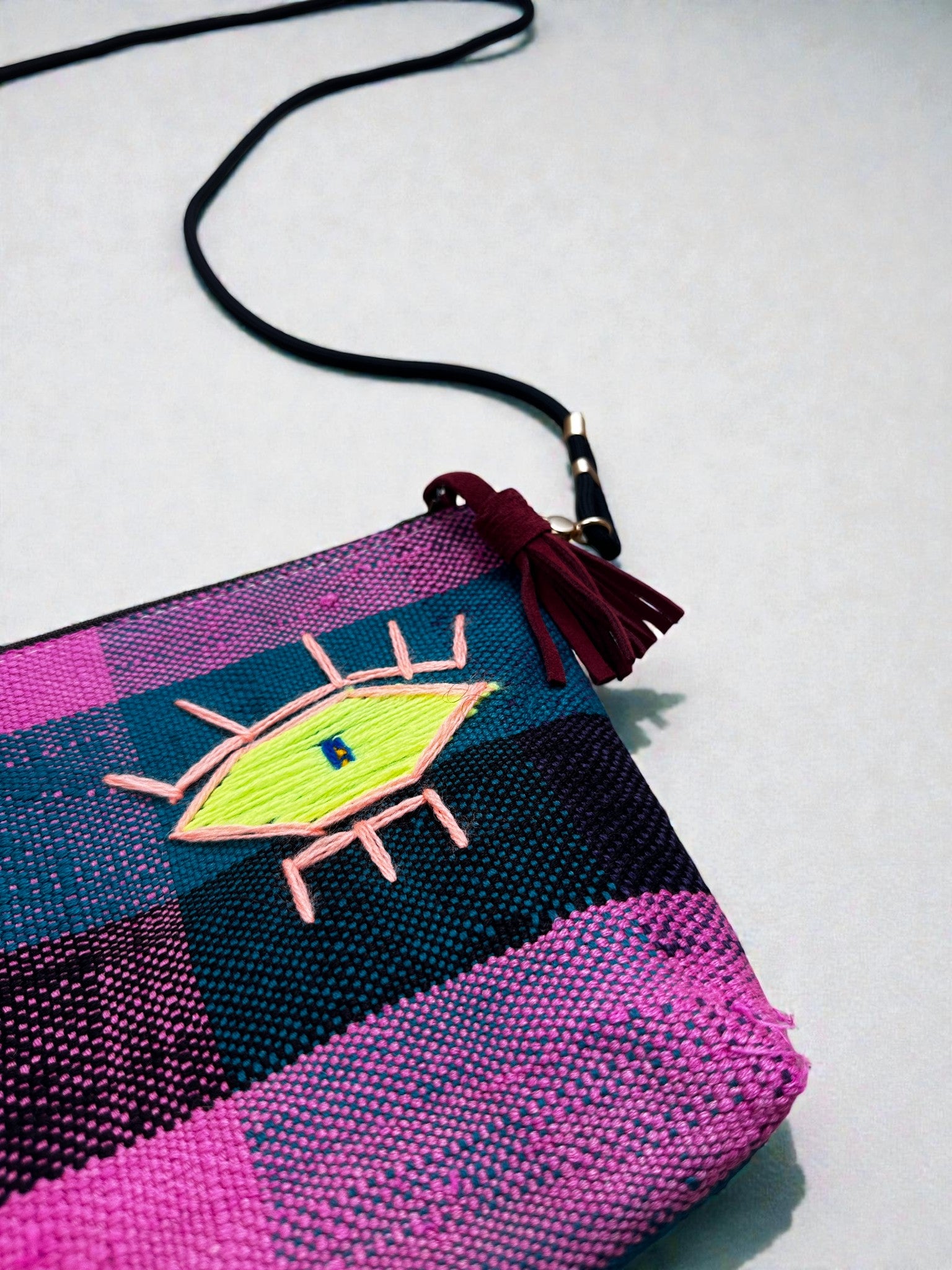 Die LEY! Tasche - OCULIA CLUTCH von LEY! ist eine karierte, handgefertigte Umhängetasche in Rosa, Lila und Blau mit einem gestickten gelben Auge und einer bordeauxfarbenen Quaste an einem schwarzen Riemen, die auf einer hellen Oberfläche ausgestellt ist.