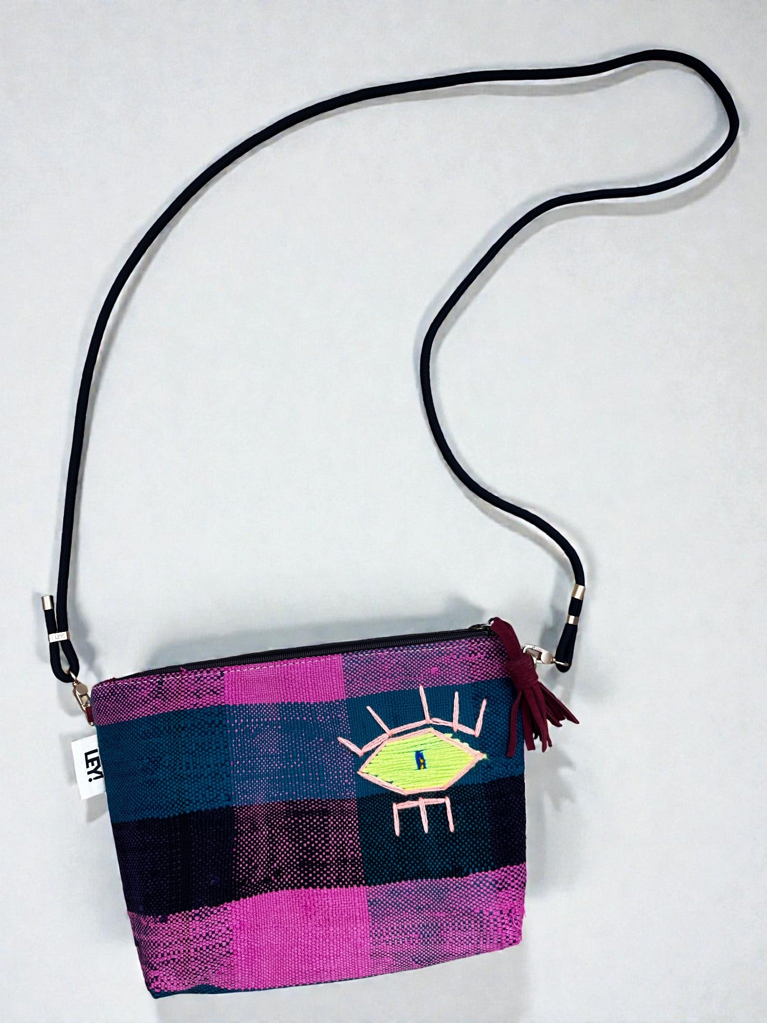 Die LEY! Tasche - OCULIA CLUTCH von LEY! ist eine kleine rechteckige Tasche aus rosafarbenem und blauem Vintage-Stoff, die mit einer gestickten Öse, einem langen schwarzen Riemen und einer roten Reißverschlussquaste ausgestattet ist - die perfekte handgefertigte Umhängetasche.