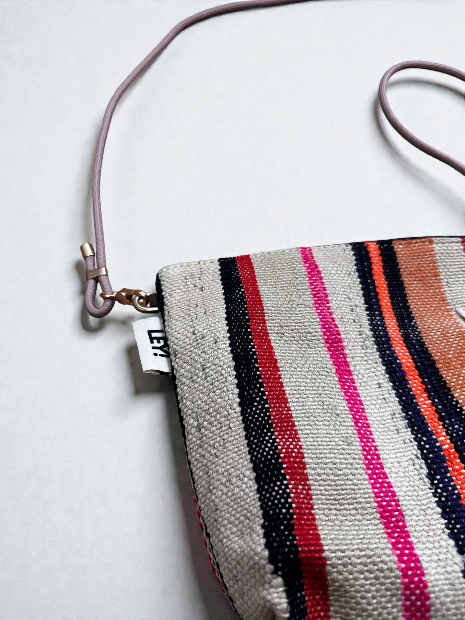 Eine Nahaufnahme der LEY! Tasche - OCULIA CLUTCH von LEY!, einer handgefertigten Umhängetasche mit rosa, orangen, schwarzen und beigen Streifen, einem hellvioletten Riemen und einem kleinen weißen Anhänger in der Nähe des Verschlusses.