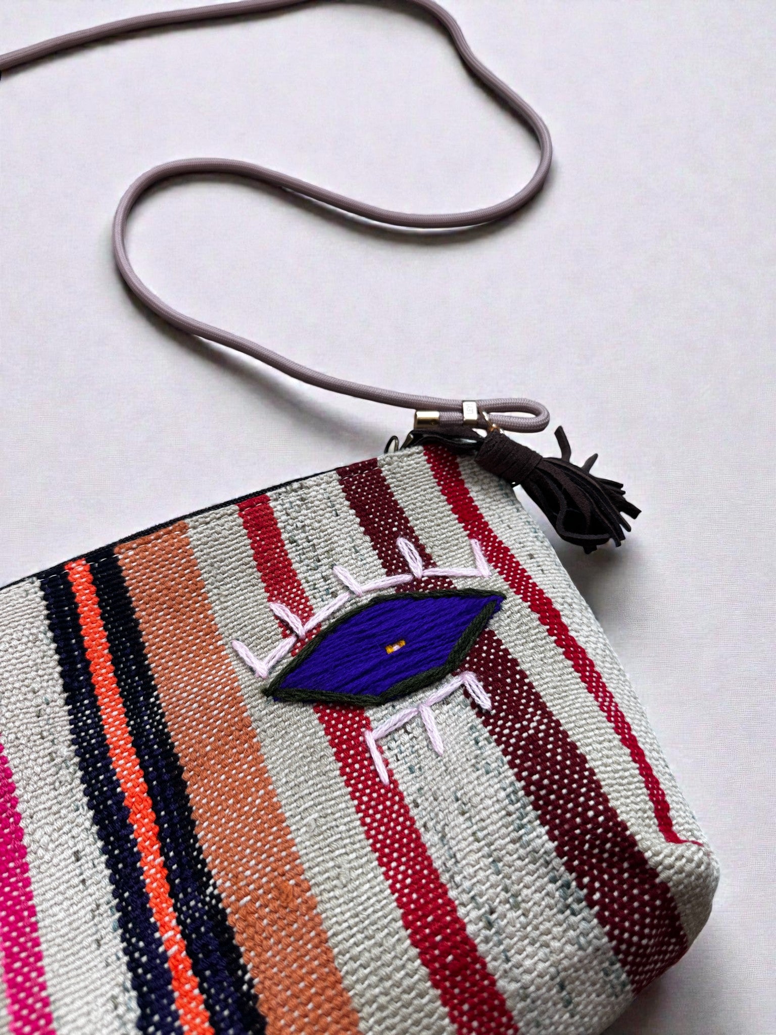 Die LEY! Tasche - OCULIA CLUTCH von LEY! ist eine farbenfrohe, gewebte Clutch mit vertikalen Streifen in Rot, Orange, Pink und Marineblau, einem gestickten blauen Auge mit weißen Wimpern und einem Reißverschluss mit Quaste auf hellem Grund.