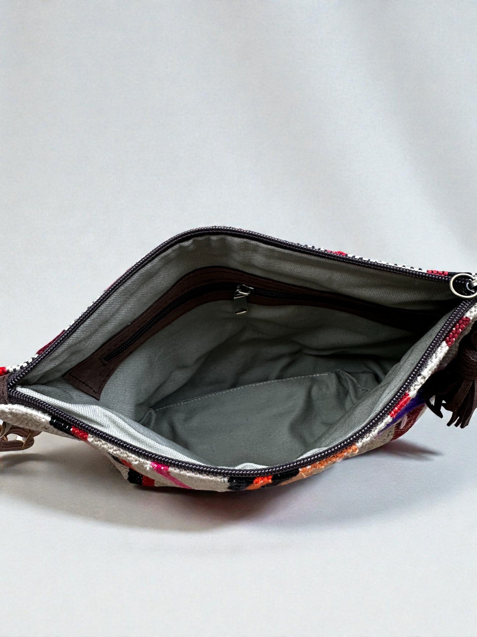 Die LEY! Tasche - OCULIA CLUTCH von LEY! ist eine kleine, handgefertigte Umhängetasche mit bunt gemusterter Außenseite, grauem Innenfutter und Reißverschluss-Innentasche, die geöffnet auf einem schlichten hellen Hintergrund abgebildet ist.
