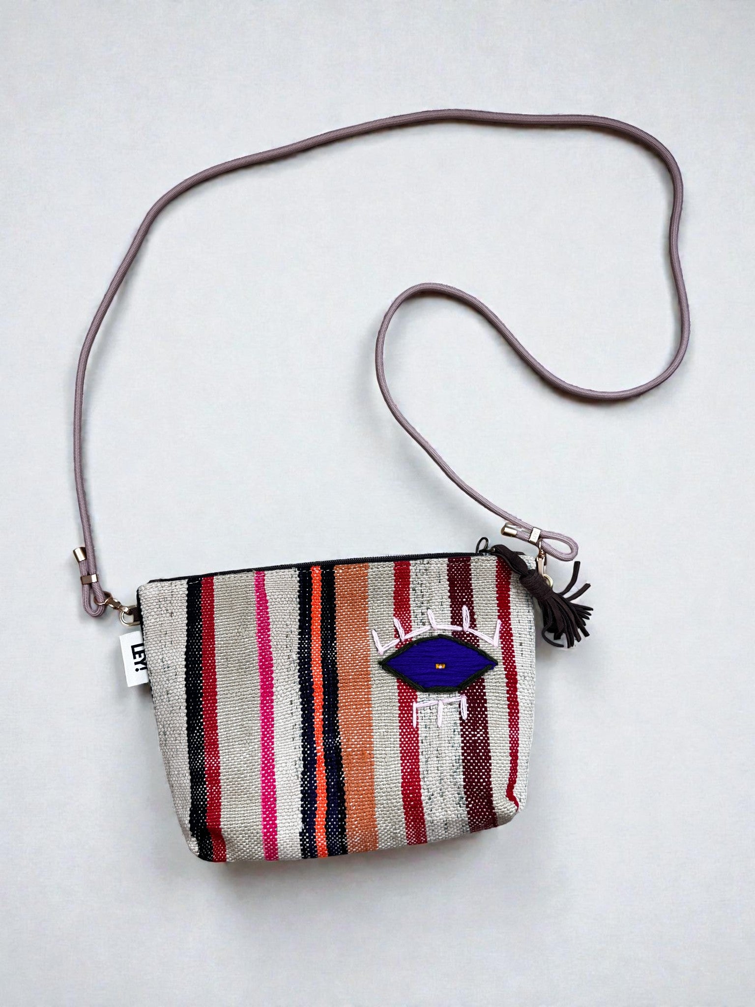 Die LEY! Tasche - OCULIA CLUTCH von LEY! ist eine kompakte, handgefertigte Crossbody-Tasche mit lilafarbenem Trageriemen, mehrfarbig gestreiftem Korpus (pink, rot, orange, grau), blauer Augenklappe und schwarzem Reißverschluss mit Quaste.