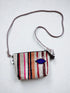 Die LEY! Tasche - OCULIA CLUTCH von LEY! ist eine kompakte, handgefertigte Crossbody-Tasche mit lilafarbenem Trageriemen, mehrfarbig gestreiftem Korpus (pink, rot, orange, grau), blauer Augenklappe und schwarzem Reißverschluss mit Quaste.