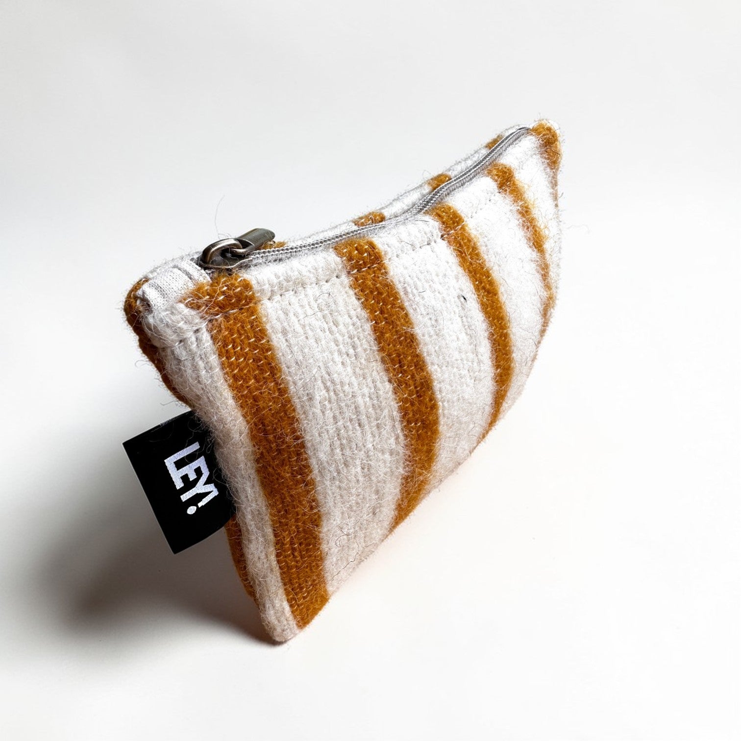 Die LEY! Tasche - WINTER EDITION - SONNE (15 x 20 cm) ist eine kleine Reißverschlusstasche aus Schurwolle in zartem weiß und senfgelb gestreift. Mit einem schwarzen "LEY!"-Anhänger an der Seite. Fotografiert vor einem schlichten weißen Hintergrund.