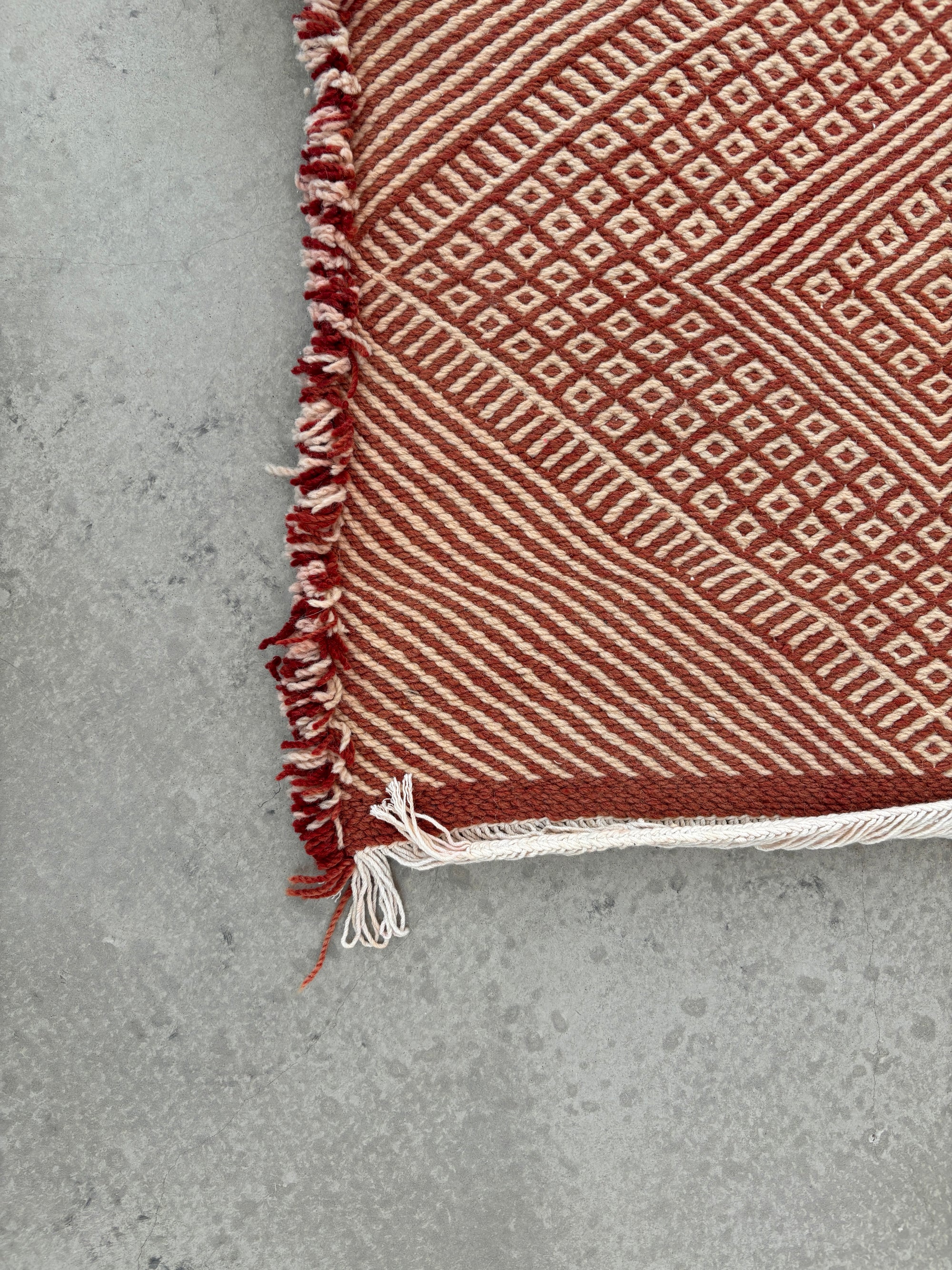 Eine Nahaufnahme des LEY-Teppichs "Sonia" von LEY! mit seinen roten und weißen geometrischen Mustern und ausgefransten Rändern zeigt seinen handwerklichen Charme auf einem glatten grauen Betonboden. Größe: 1,95 x 3,00 m.