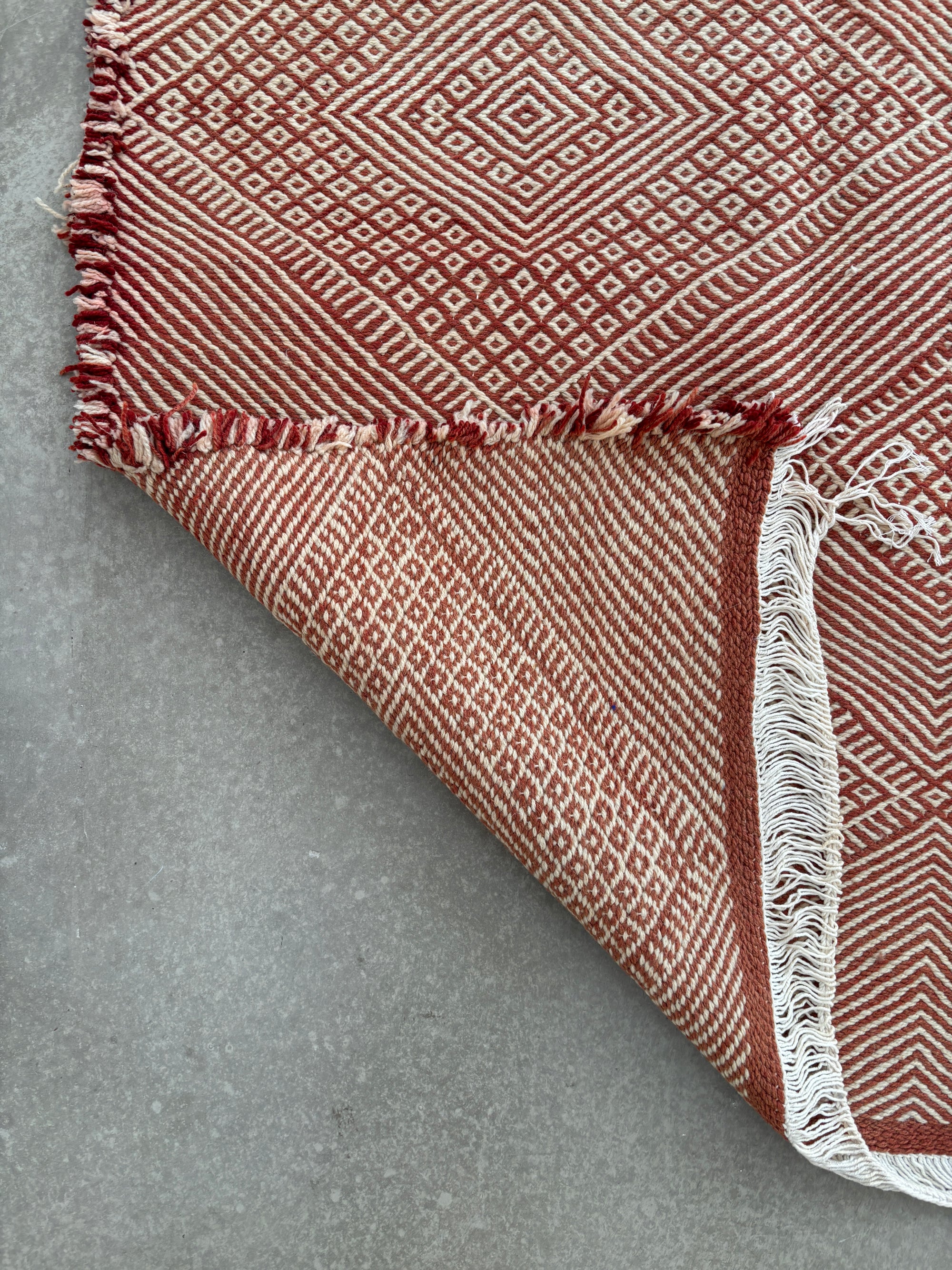 Eine Nahaufnahme des LEY-Teppichs "Sonia" 1,95 x 3,00 von LEY! mit roten und weißen geometrischen Mustern, ausgefransten Rändern und einer gefalteten Ecke, dargestellt auf einer grauen Oberfläche.