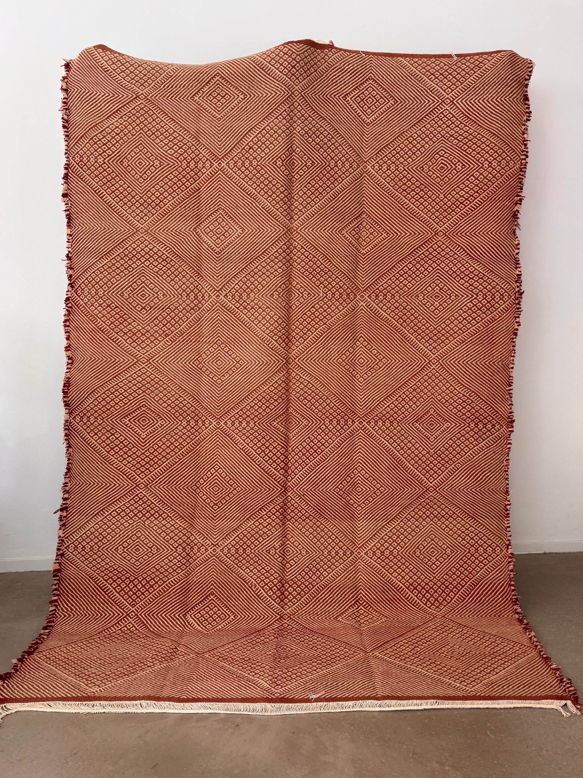 Der handgeknüpfte LEY Teppich "Sonia" (1,95 x 3,00) von LEY! zeigt feine rote und beige Diamantmuster, ist mit Fransen an den Seiten versehen und wird vertikal an einer weißen Wand präsentiert.