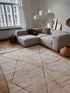Ein modernes Wohnzimmer mit einer großen beigen Couchgarnitur, dem handgeknüpften BERBER TEPPICH "ILAJHA" IM WUNSCHMAß von LEY!, Weidenkörben, gerahmter Kunst, weißen Pendelleuchten und Sonnenlicht, das durch hohe Fenster mit weißen Vorhängen fällt.