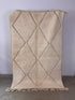 Der BERBER TEPPICH "JUNAH" von LEY! (1,50 x 2,50 m) hat eine beigefarbene, strukturierte Oberfläche mit einem einfachen Rautenmuster und fransigen Quasten am unteren Rand, die aufrecht an einer hellen Wand angebracht sind.
