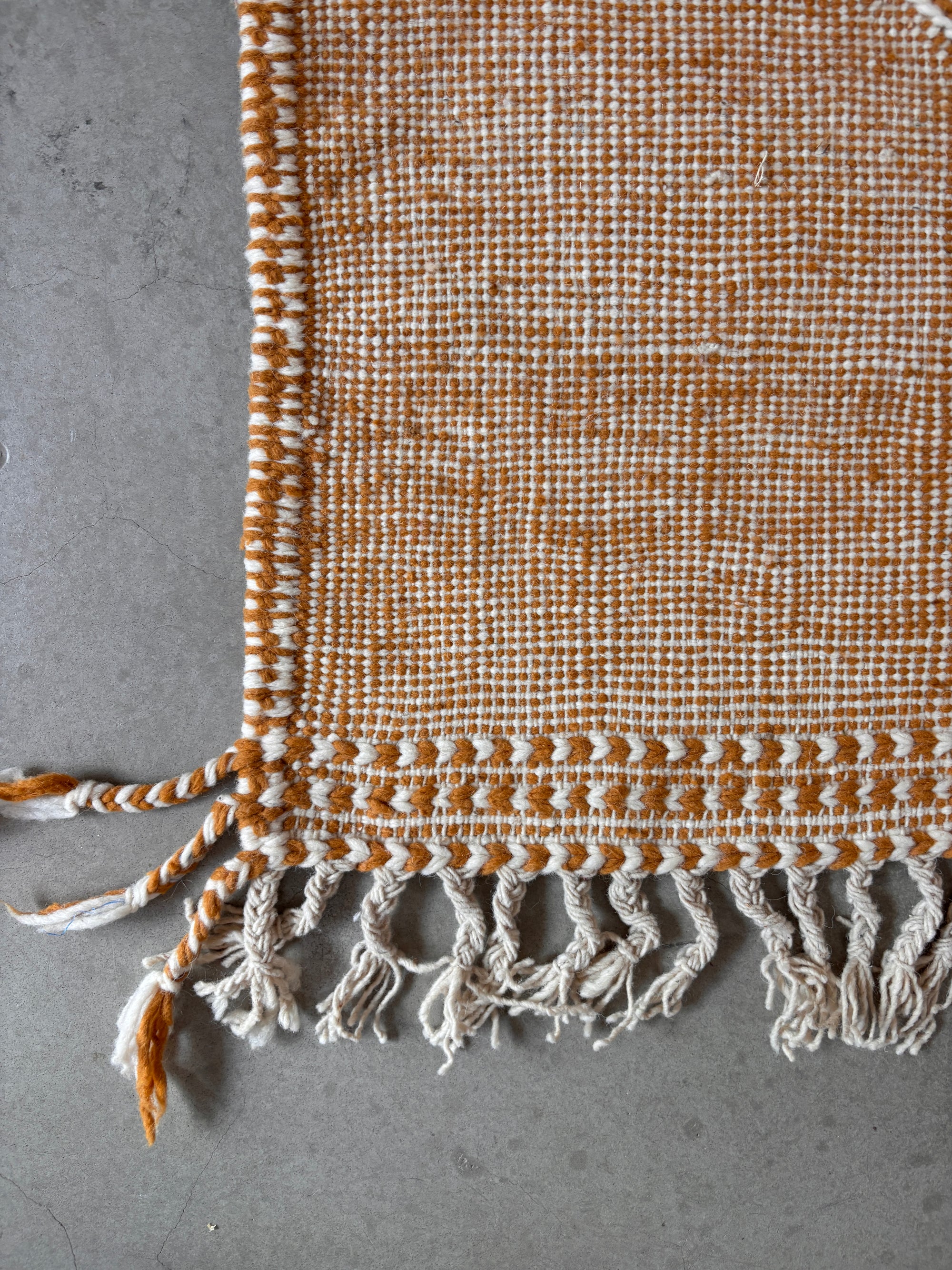 Eine Nahaufnahme des LEY Teppich - Kastira (1,50 x 2,55) von LEY! mit braun-weißem Karomuster, gedrehten Fransen und weißen Quasten auf einem grauen Steinboden.