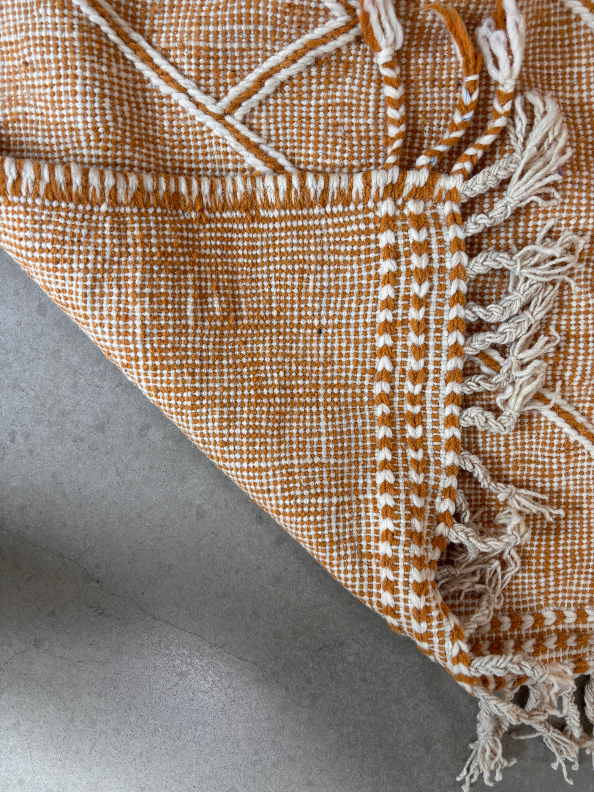 Eine Nahaufnahme des LEY Teppich - Kastira (1,50 x 2,55) von LEY!, ein orange-weißer handgeknüpfter Kelim mit geometrischem Muster und ausgefransten Kanten, teilweise gefaltet auf einer glatten grauen Oberfläche.