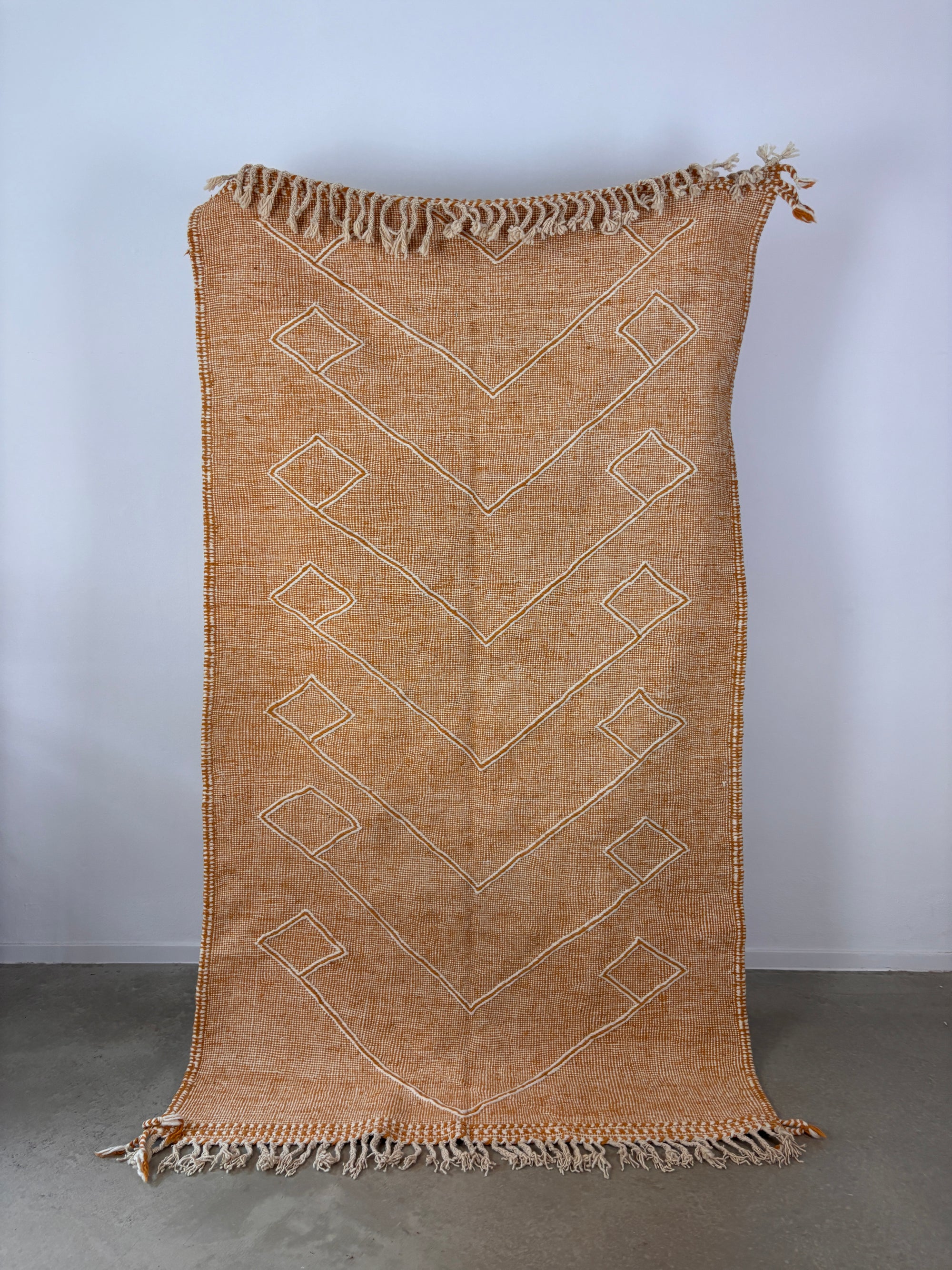 Der LEY! LEY Teppich - Kastira - 1,50 x 2,55 ist ein rechteckiger, hellbrauner Flachgewebe-Teppich mit weißen Chevron- und Rautenmustern und ausgefransten Kanten, der aufrecht vor einer hellgrauen Wand auf einem Betonboden gezeigt wird.