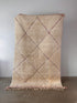 Der beige LEY! Berber Teppich - Perla (1,50 x 2,50) aus 100 % Schurwolle mit schlichtem Rautenmuster in dünnen rosa Linien steht auf Betonboden vor grauer Wand und hat Fransen am unteren Rand.