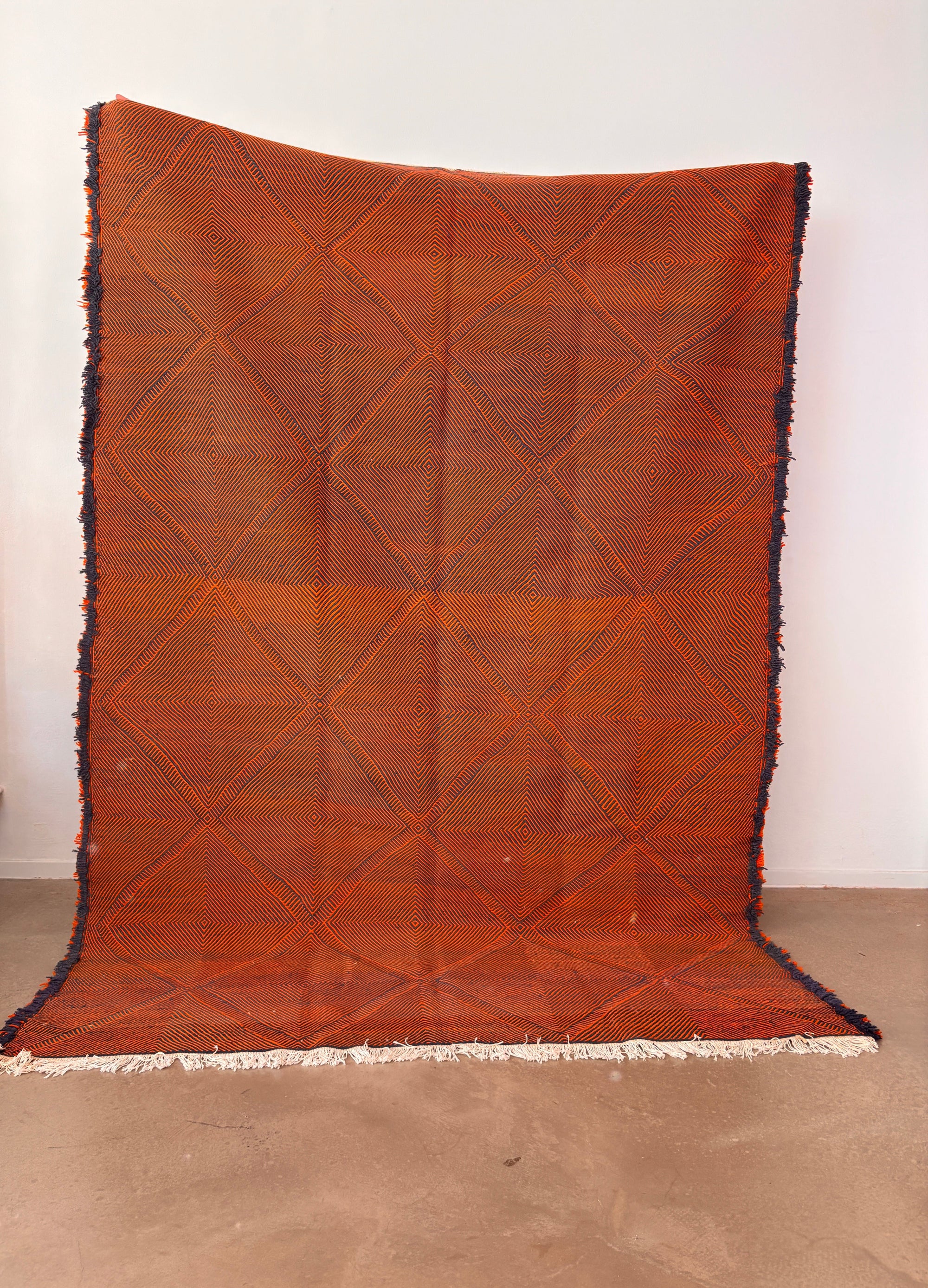 Der LEY Teppich "Rabina" 2,00 x 3,00 von LEY! ist ein großer, orangefarbener Flachgewebe-Teppich mit dezentem Rautenmuster und ausgefransten Rändern, der aufrecht vor einer schlichten weißen Wand auf einem glatten braunen Boden gezeigt wird.