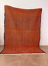 Der LEY Teppich "Rabina" 2,00 x 3,00 von LEY! ist ein großer, orangefarbener Flachgewebe-Teppich mit dezentem Rautenmuster und ausgefransten Rändern, der aufrecht vor einer schlichten weißen Wand auf einem glatten braunen Boden gezeigt wird.