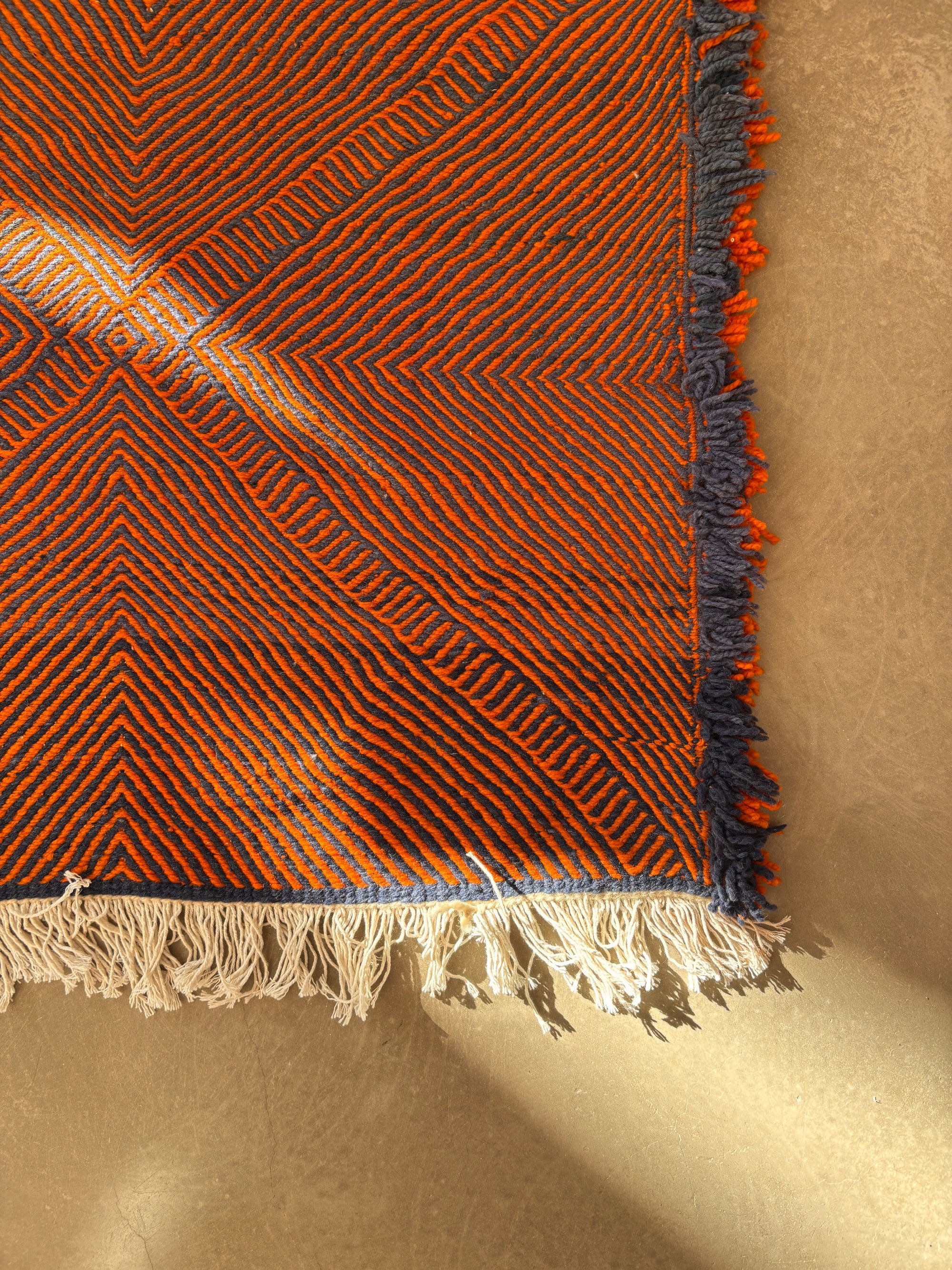 Eine Nahaufnahme des LEY-Teppichs "Rabina" 2,00 x 3,00 von LEY! mit orangefarbenen und dunkelblauen Zickzack-Mustern, blauen und weißen Fransen am Rand, wobei das Sonnenlicht Schatten auf einen hellbraunen Boden wirft.