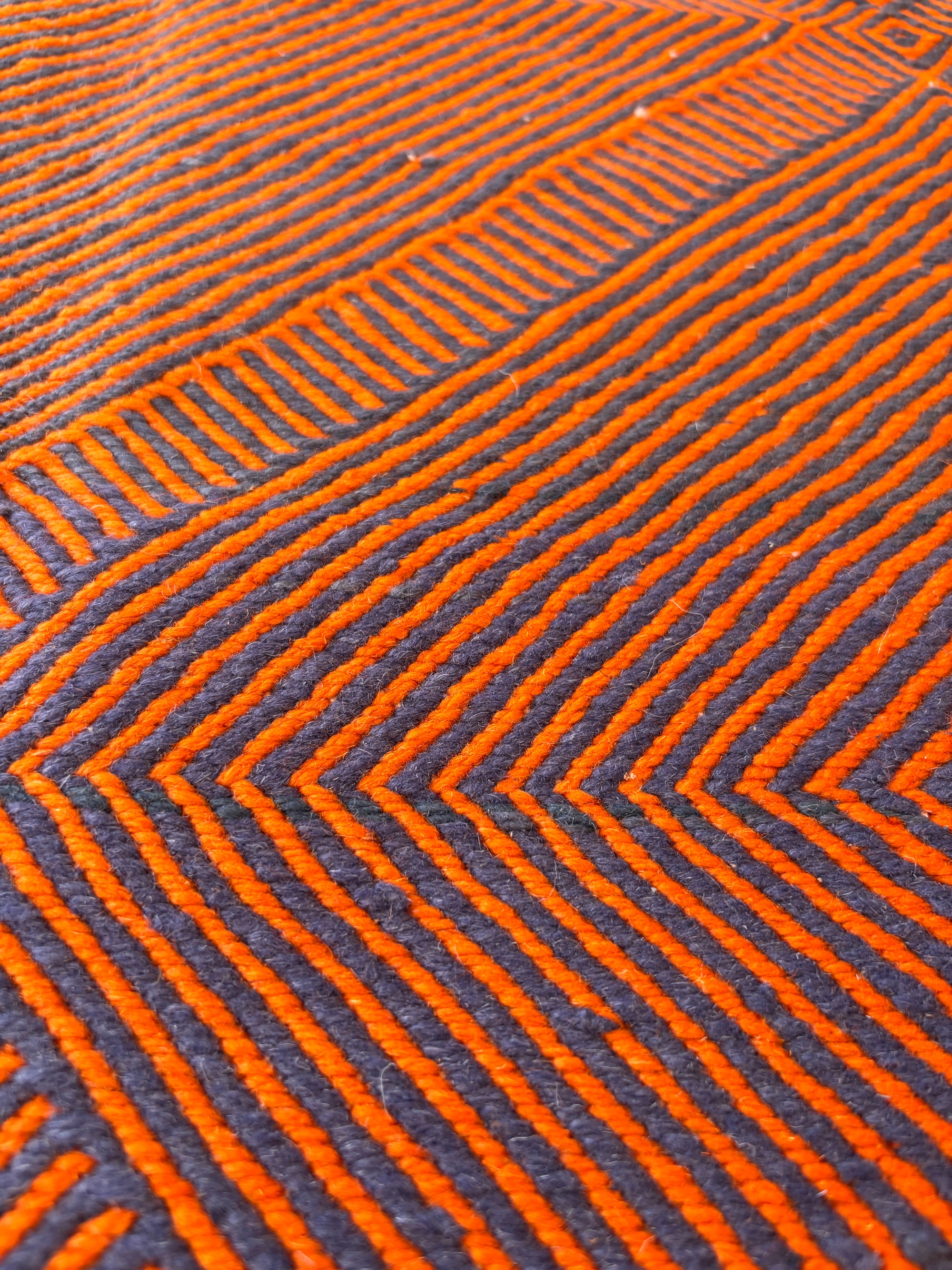 Nahaufnahme des LEY Teppichs "Rabina" 2,00 x 3,00 von LEY! mit parallelen orangefarbenen Chevrons auf dunkelgrauem Grund. Das geometrische, flachgewebte Design erinnert an den Teppich Marokko und bringt lebendige Tiefe und Bewegung in Ihren Raum.