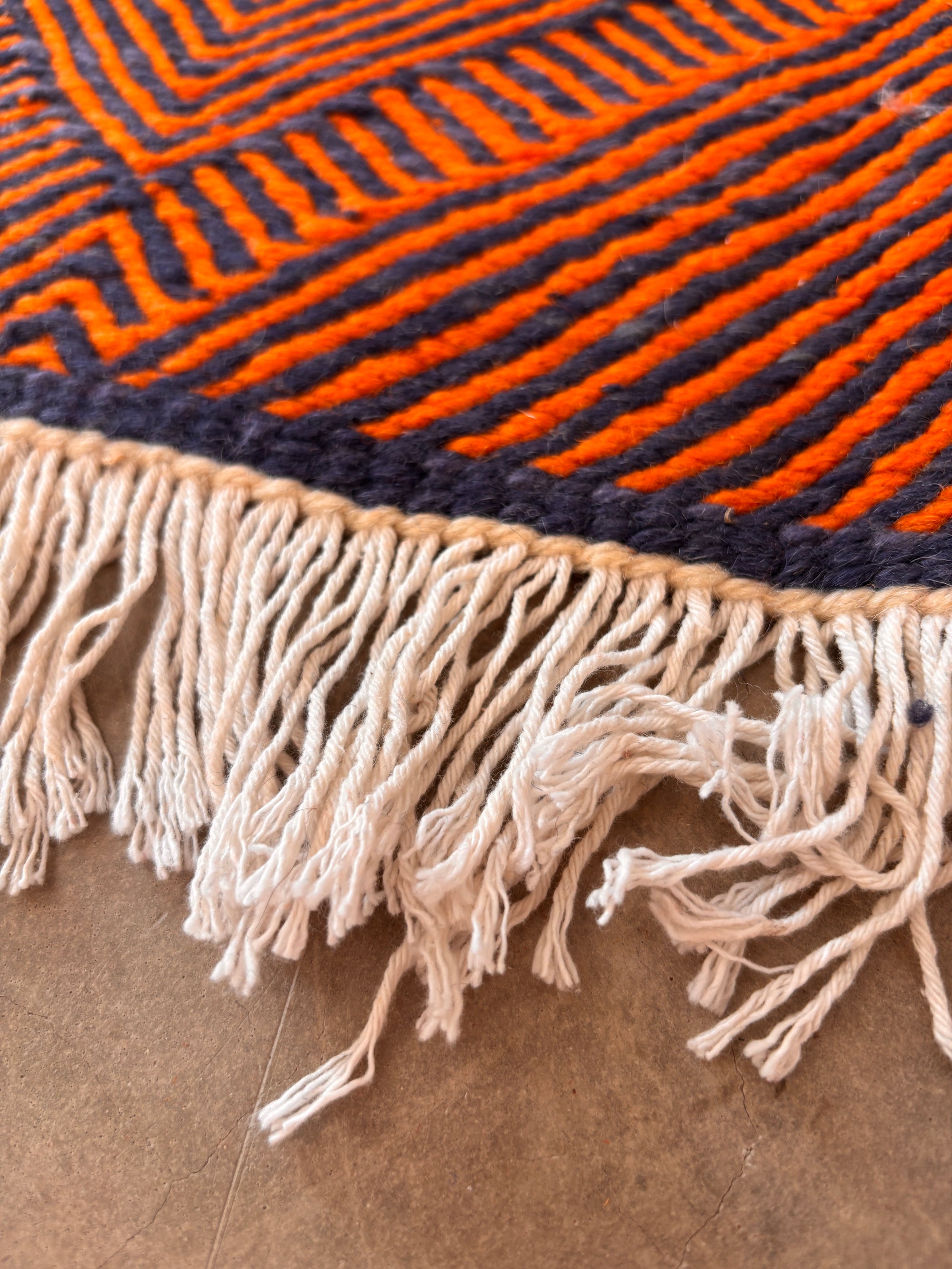 Nahaufnahme des LEY Teppichs "Rabina" 2,00 x 3,00 von LEY! mit orangefarbenen und marineblauen geometrischen Streifen und weißen Fransen, verlegt auf einem beigen Boden. Dieser flachgewebte Teppich bringt handwerklichen Marokko-Stil in Ihren Raum.