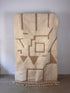 Der Berberteppich - TAZIN (1,50 x 2,50) von LEY! aus beiger und hellbrauner, handgewebter Wolle mit geometrischen und abstrakten Mustern und Fransen lehnt senkrecht an einer schlichten weißen Wand auf einem Betonboden.