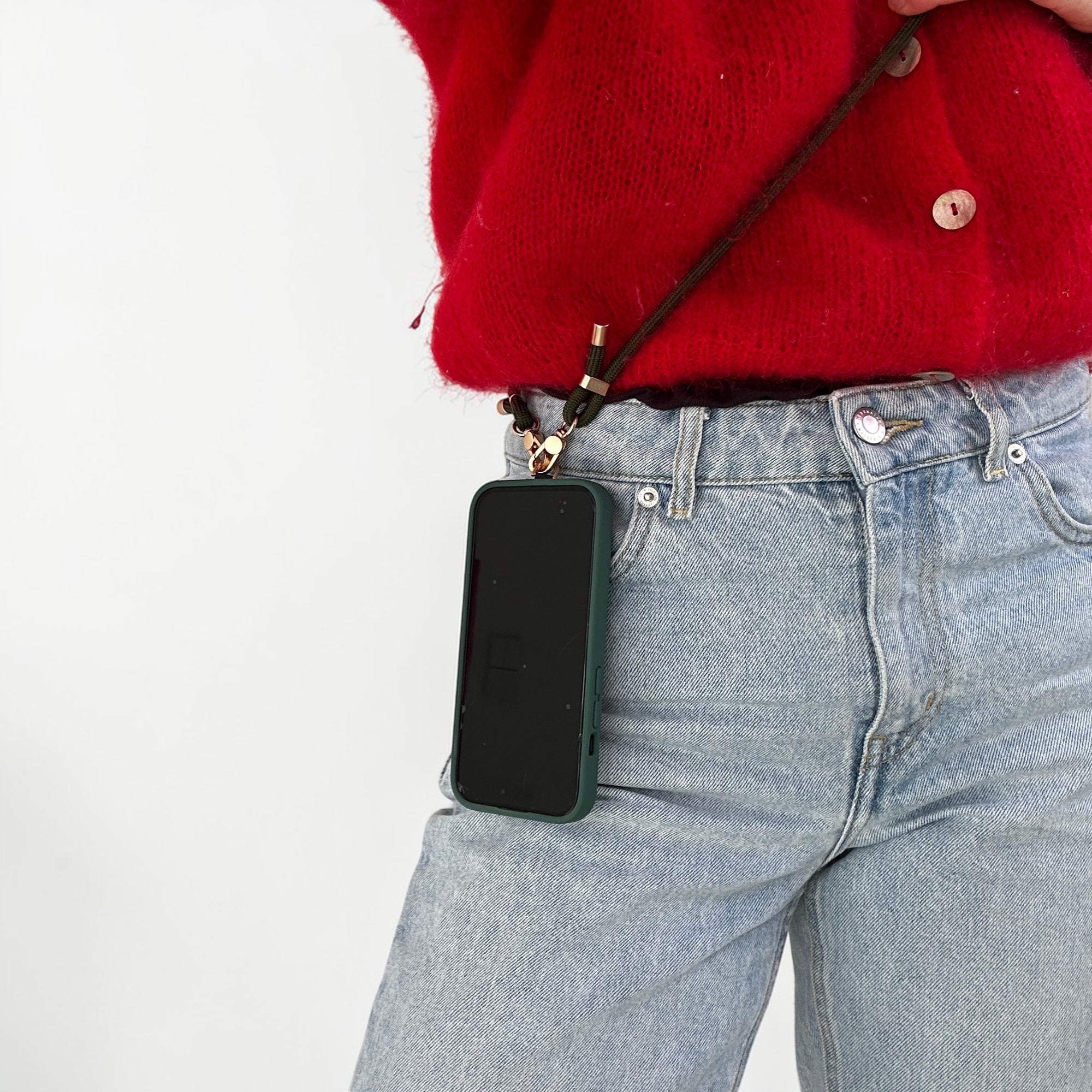 Eine Person in hellblauer Jeans und rotem Pullover trägt ein Smartphone in einer grünen Tasche mit der LEY! Handykette - Olivgrün, die an ihrer Taille hängt. Der Hintergrund ist einfarbig weiß.